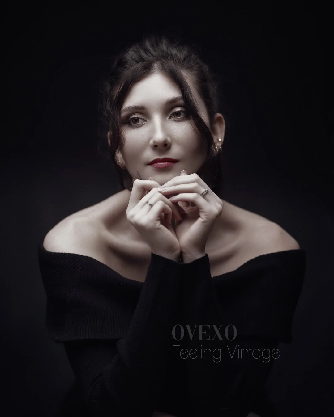 Portrait of a woman with dark hair, wearing a black off-shoulder top, against a black background, with a calm expression and her hands near her face.