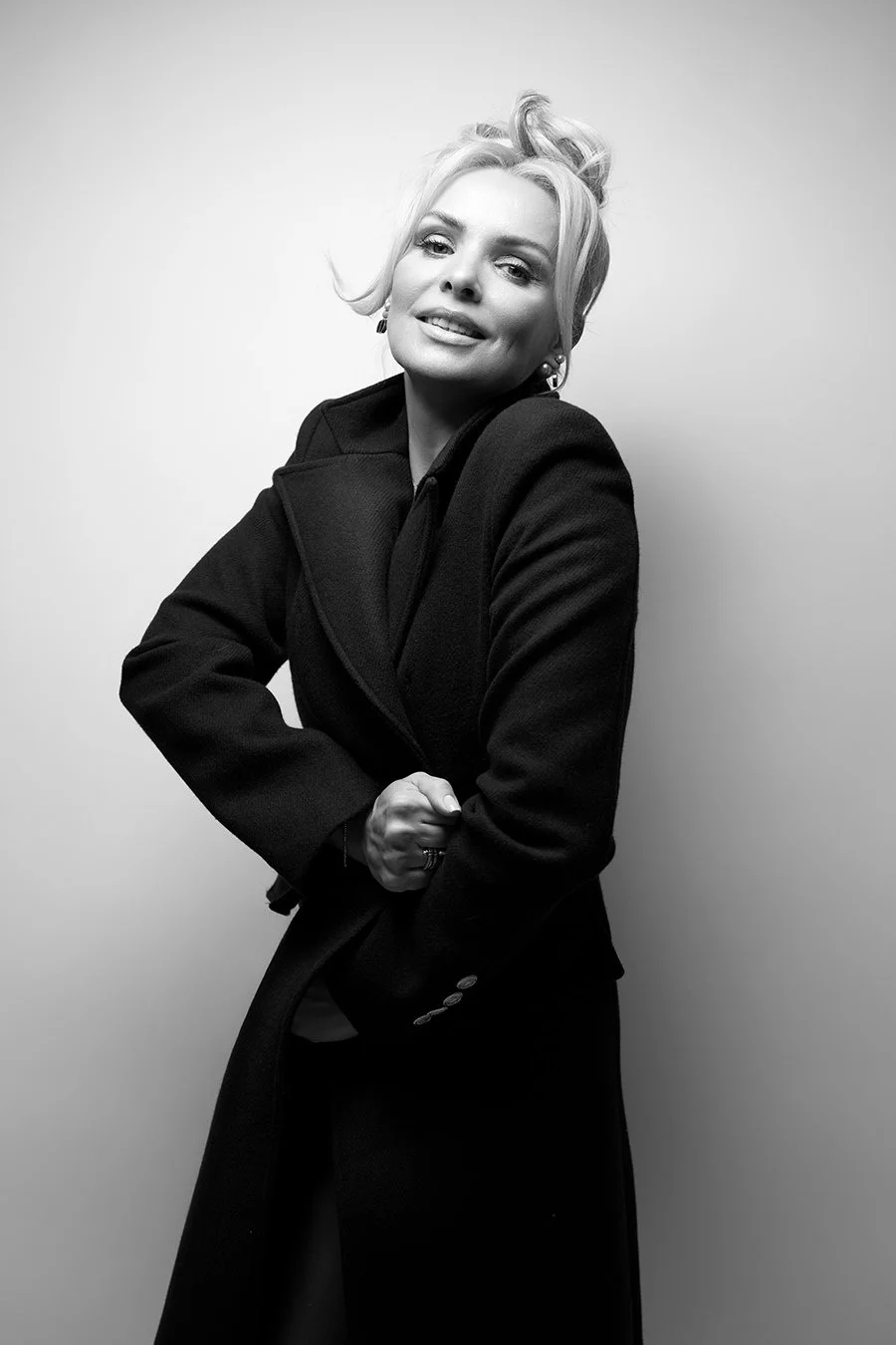 Black-and-white photo of a woman with blonde hair styled in loose waves, wearing a dark coat, smiling slightly, posing with one arm bent and the other hand resting on her waist, against a plain light background.
