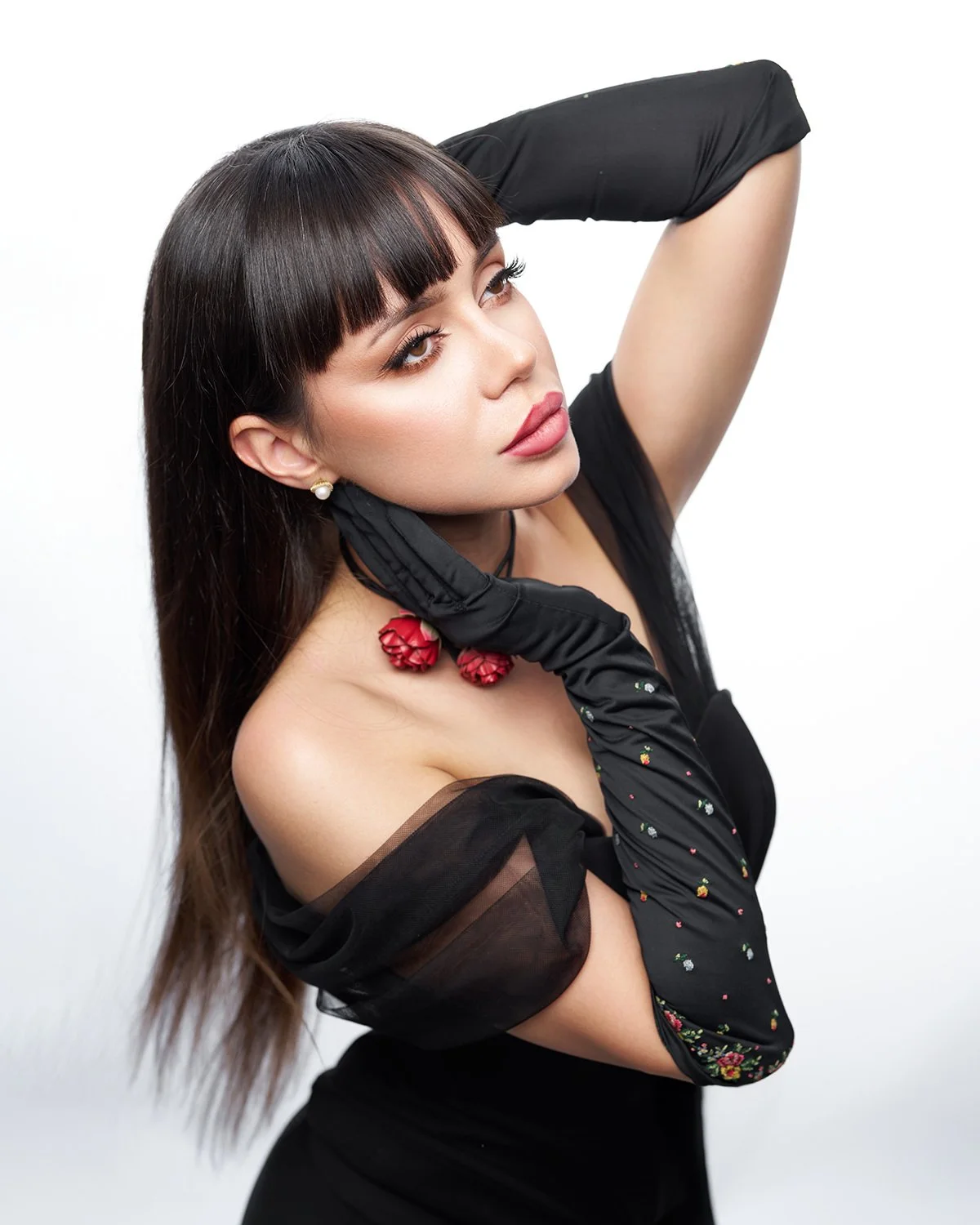 A woman with dark hair and bangs, wearing black clothing and long black gloves, is posing with one arm behind her head against a white background.