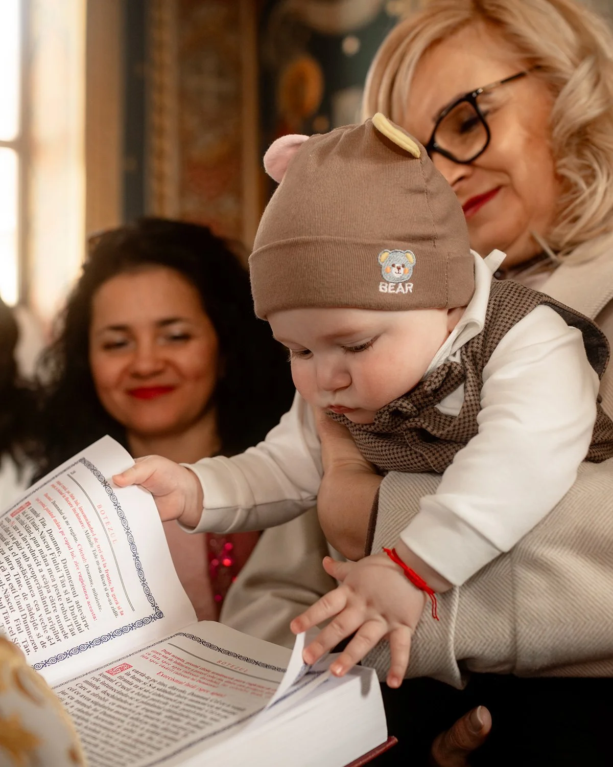 A baby in a brown bear hat looking at a book with a woman in glasses holding him, and another woman with curly black hair smiling in the background, in a cozy indoor setting.