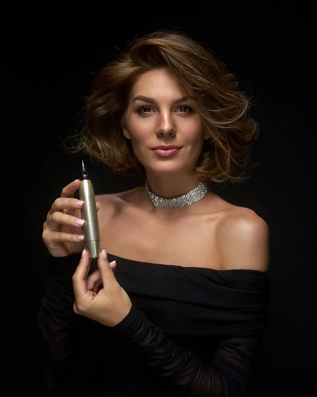 A woman with shoulder-length, wavy brown hair, wearing a black off-shoulder top, a sparkly choker necklace, and holding a silver dermal filler syringe, smiling softly against a dark background.