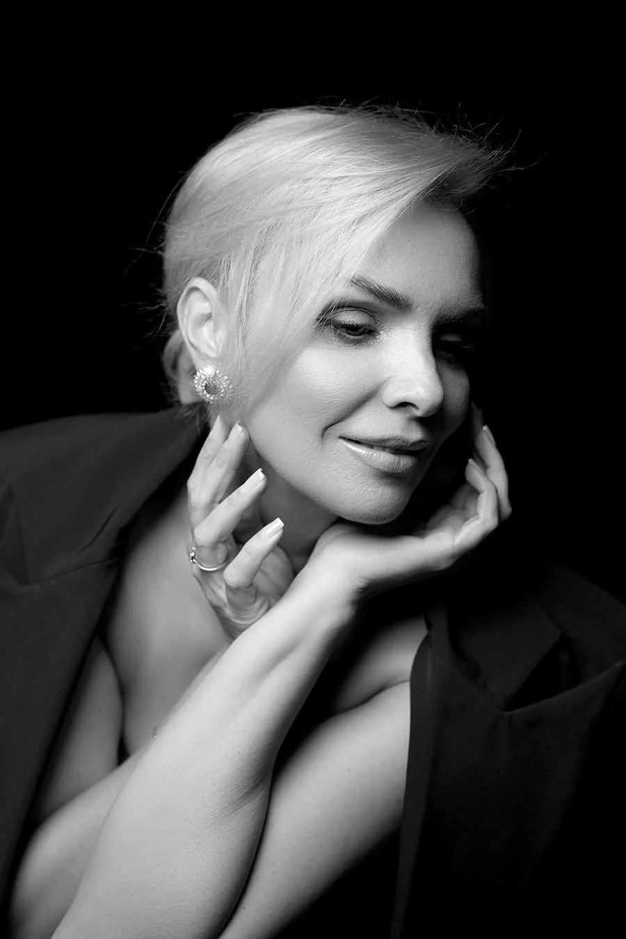 A black-and-white portrait of a woman with short blonde hair, wearing earrings and a dark blazer, with her hands gently touching her face and shoulder, smiling softly.