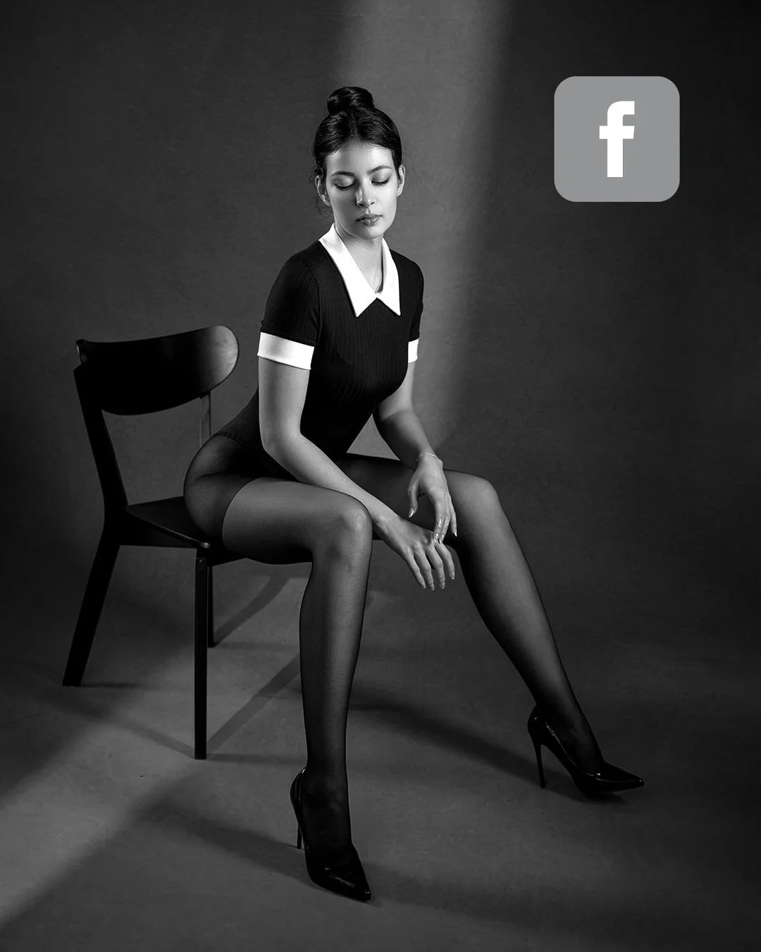 Femeie cu părul strâns în coc, purtând un body negru cu guler alb și pantofi cu toc, stând pe un scaun negru într-un studio foto alb-negru.