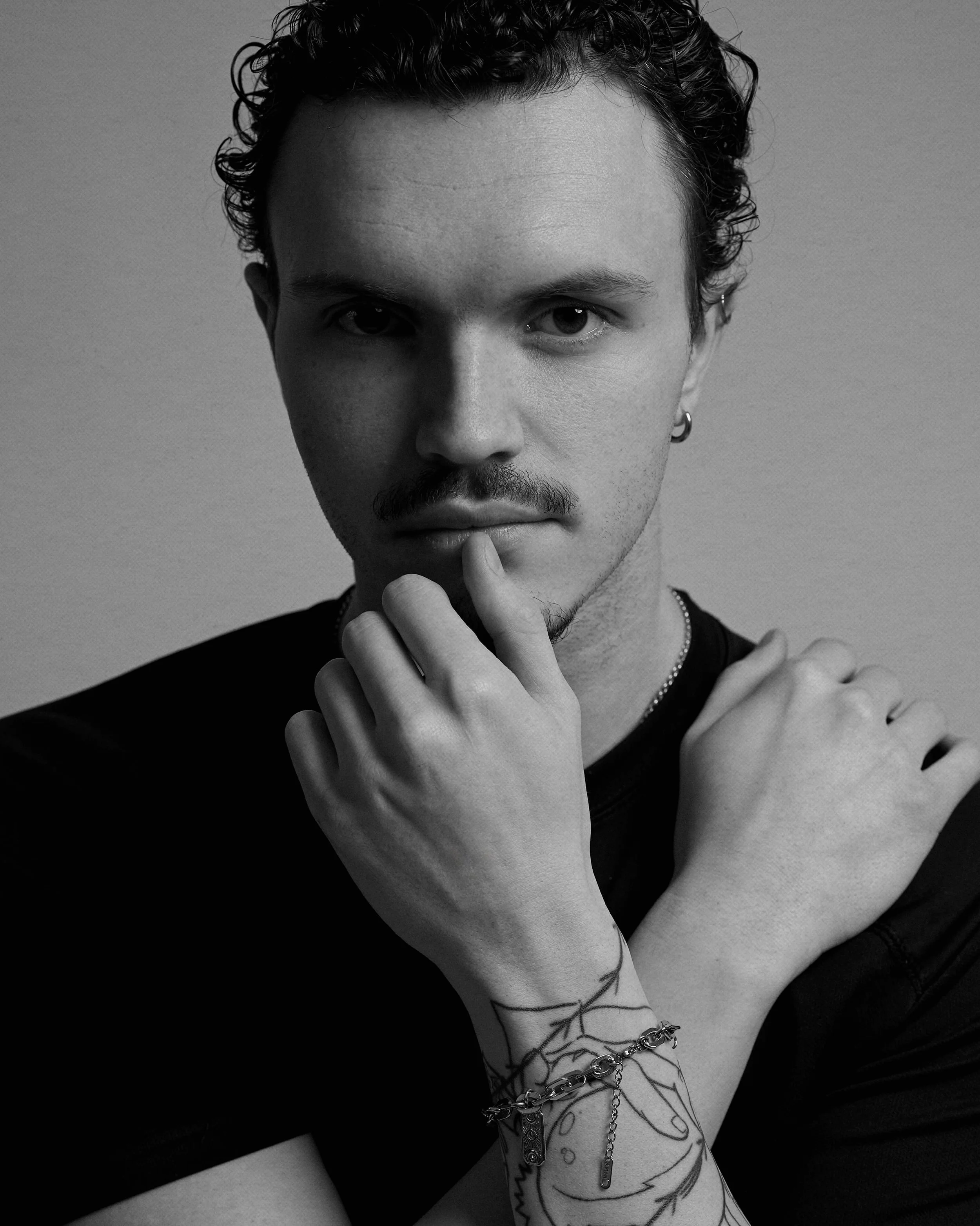 Black and white portrait of a young man with curly hair and a mustache, looking directly at the camera with a thoughtful expression, touching his chin with one hand, wearing a black shirt and jewelry, with tattoos on his arm.