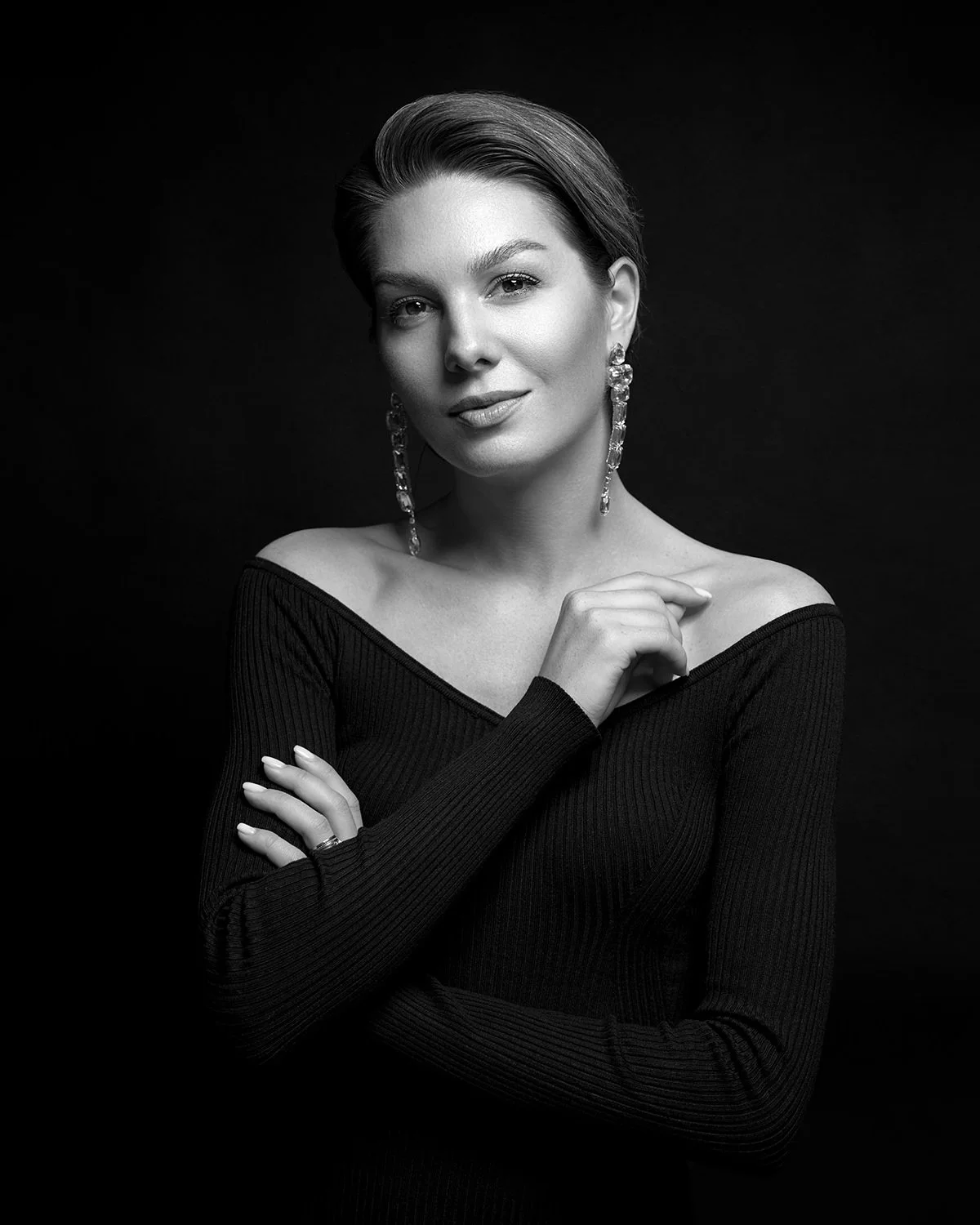 Black and white portrait of a woman with short hair, wearing earrings and a long-sleeved off-the-shoulder top, posing with her hand near her shoulder against a dark background.