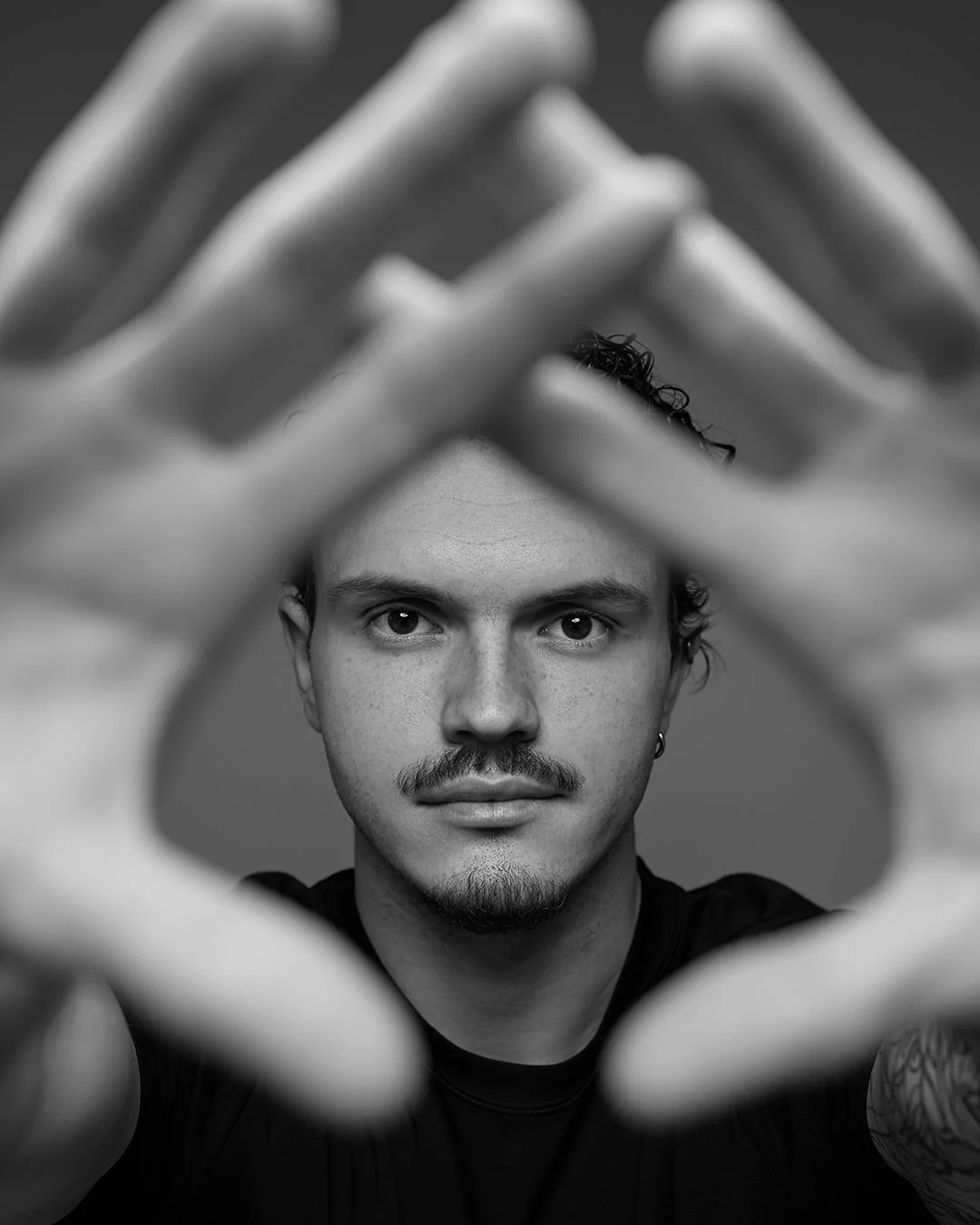 Black and white photo of a young man with short curly hair and a mustache, looking directly at the camera through a frame made by his hands.