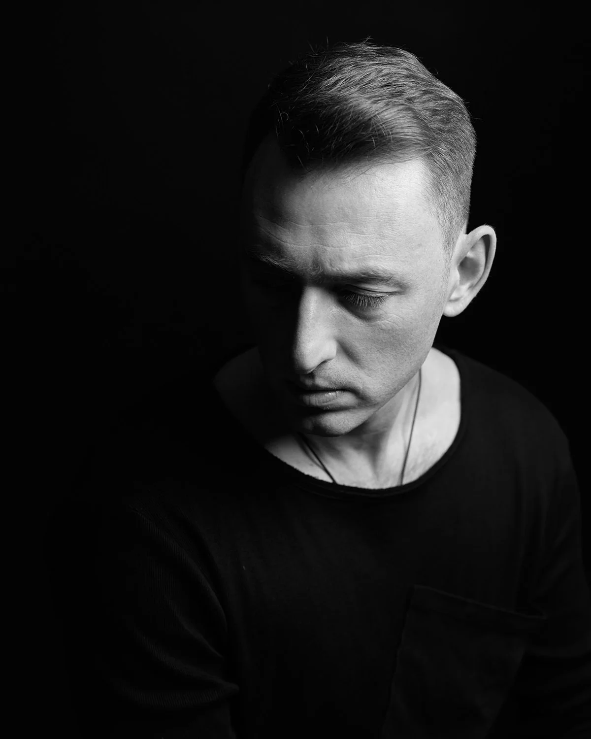 A black and white portrait of a young man with closed eyes, short hair, wearing a dark shirt and a necklace, against a dark background.