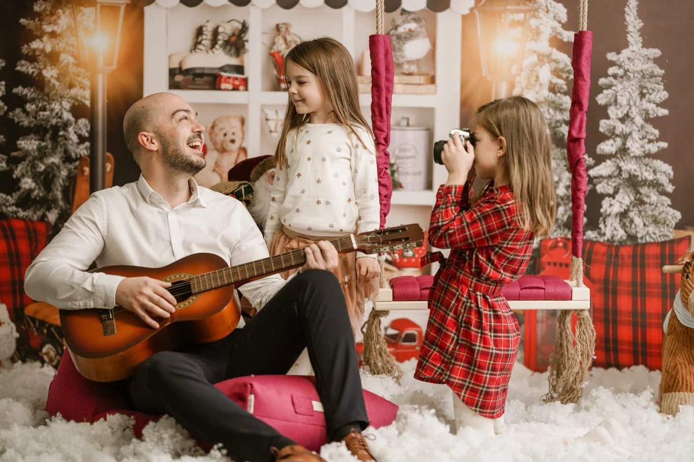 A man playing guitar and smiling at two young girls in a festive, Christmas-themed room with snow-like decorations, Christmas trees, and holiday decor