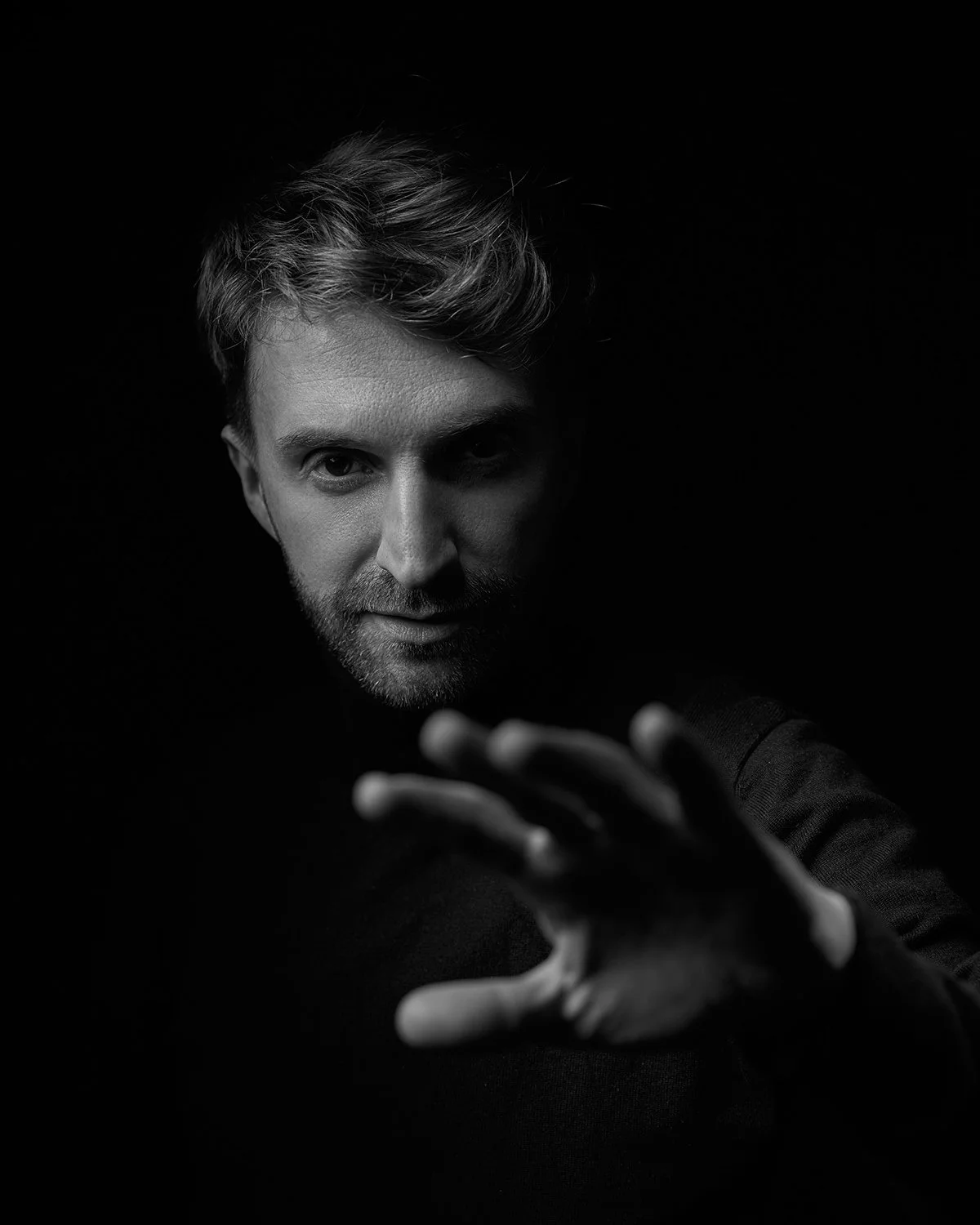 Black and white portrait of a man reaching toward the camera with a serious expression, dark background, dramatic lighting.