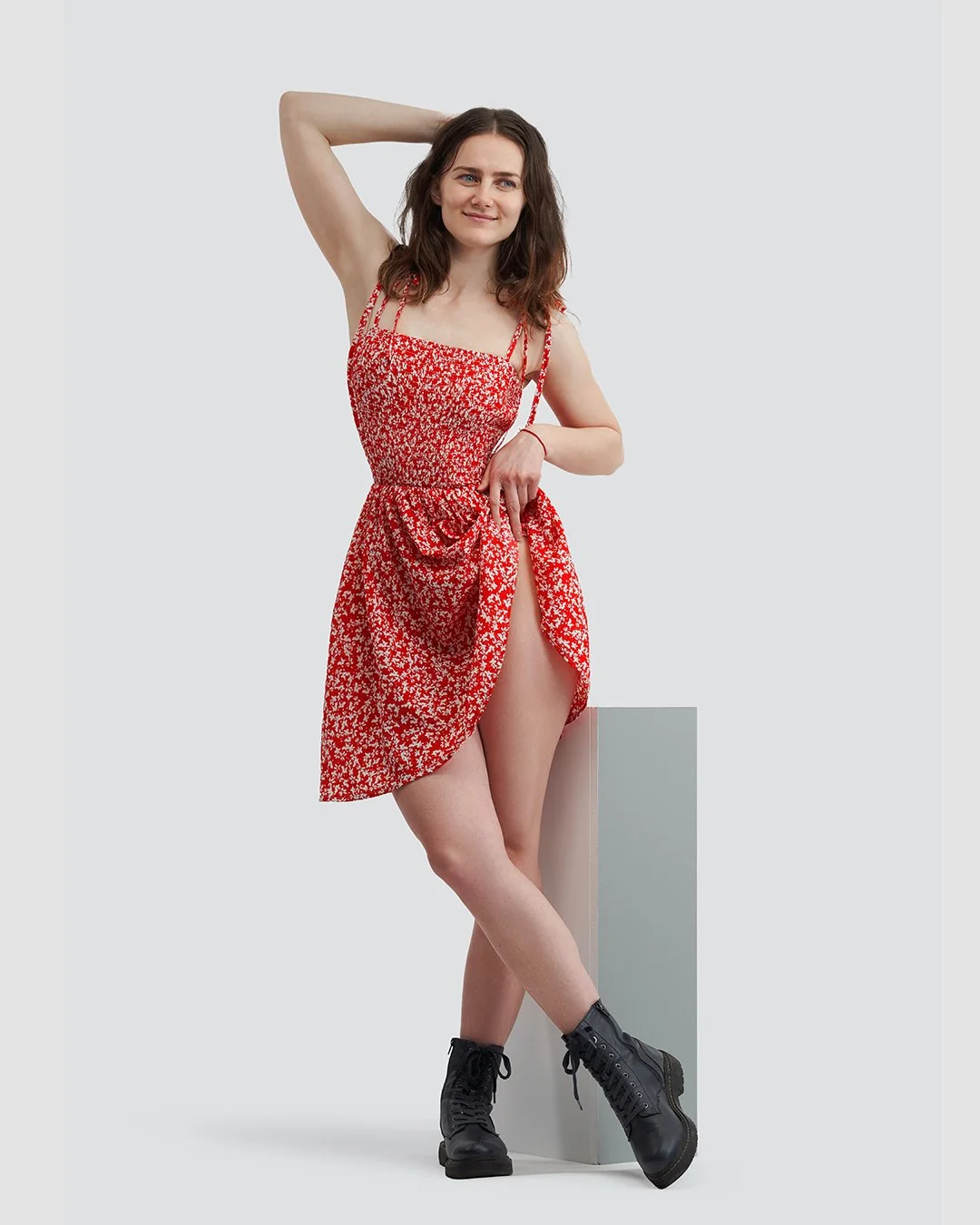 A young woman with wavy brown hair standing against a plain background, wearing a red and white floral dress with slit, black combat boots, and posing confidently with one hand behind her head and the other on her hip.
