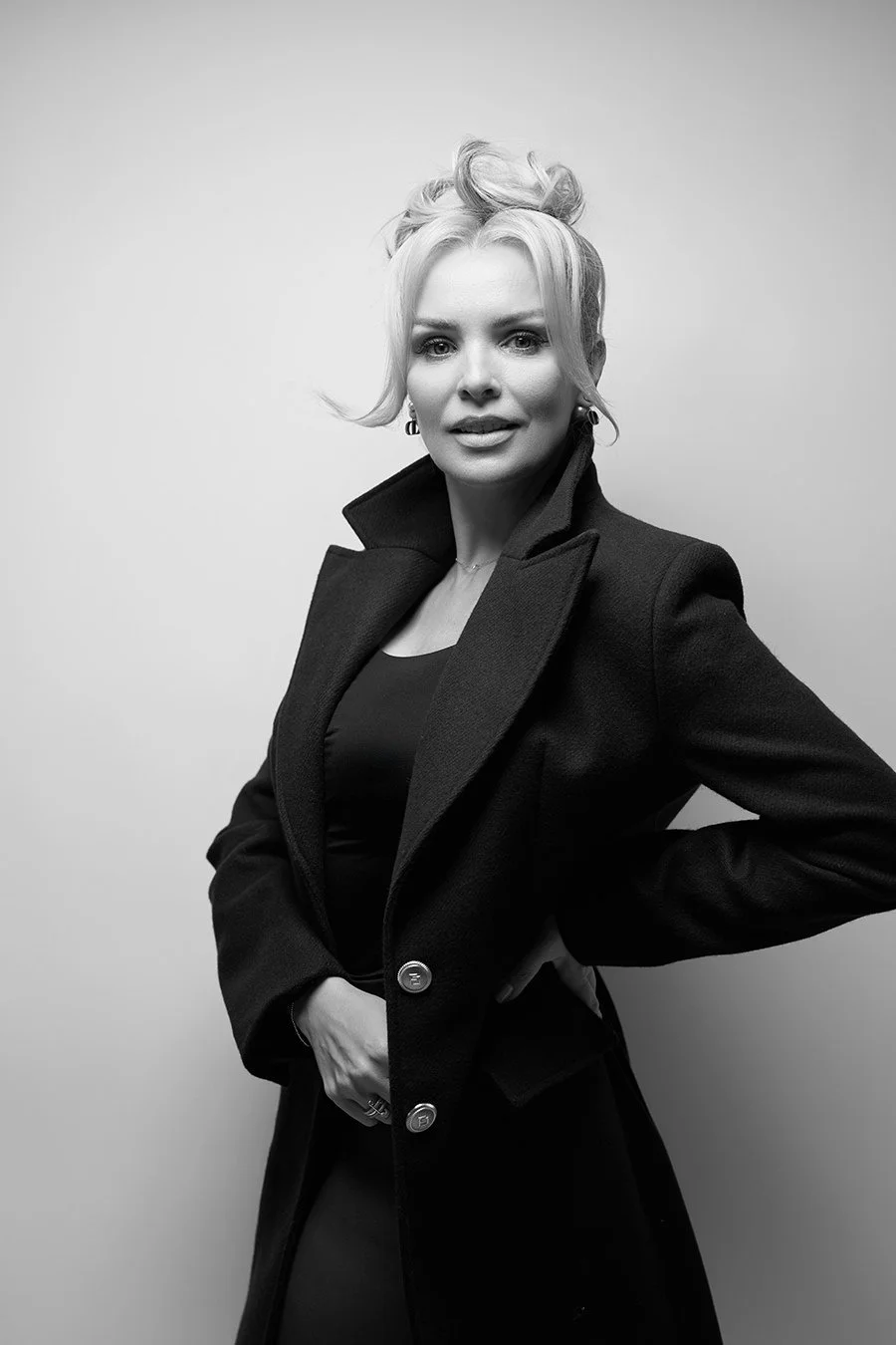 Black and white portrait of a woman with styled blonde hair, wearing a dark coat over a top, standing against a plain light background.