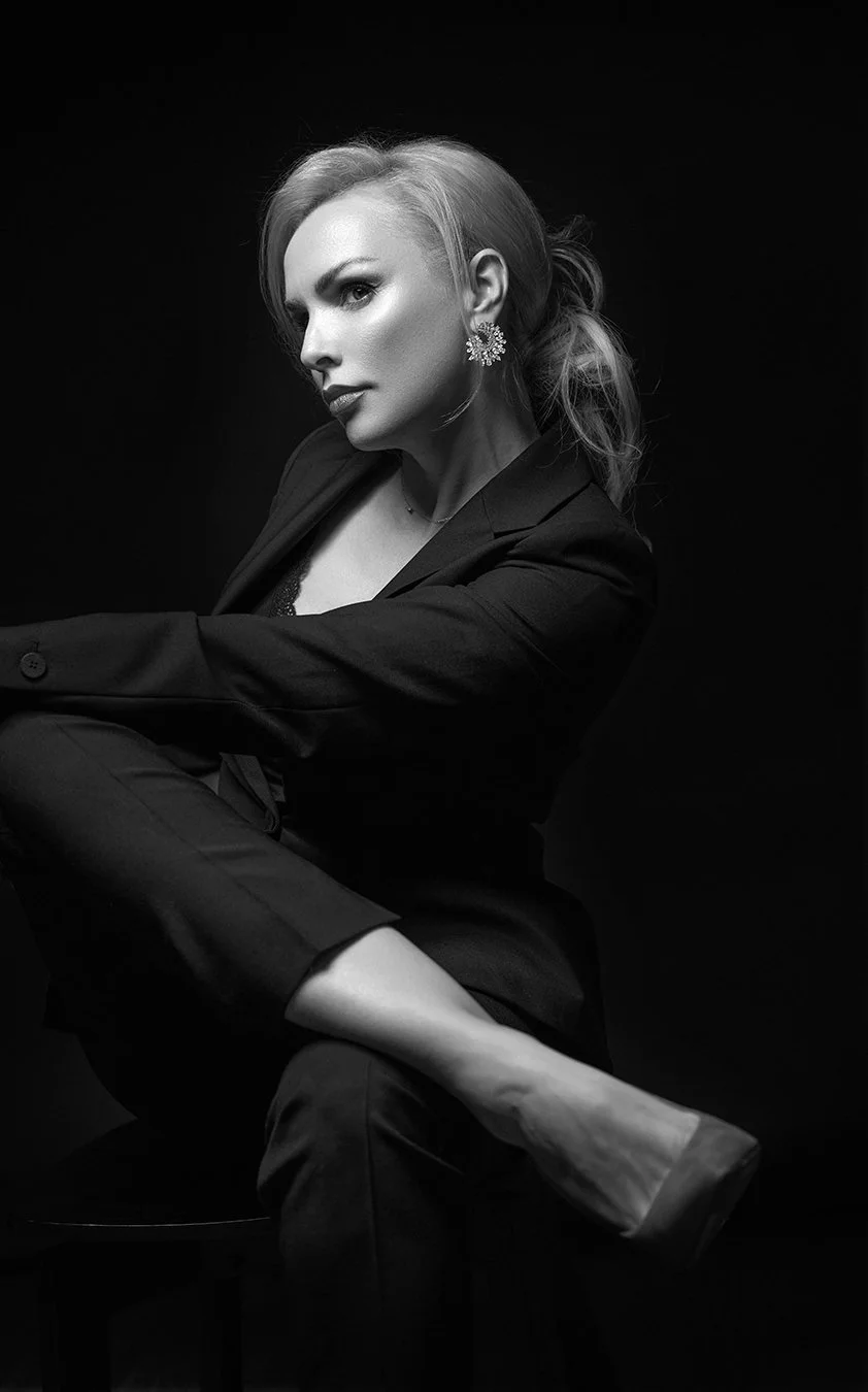 A black-and-white portrait of a woman with blonde hair, wearing a dark blazer, large earrings, and sitting against a dark background.