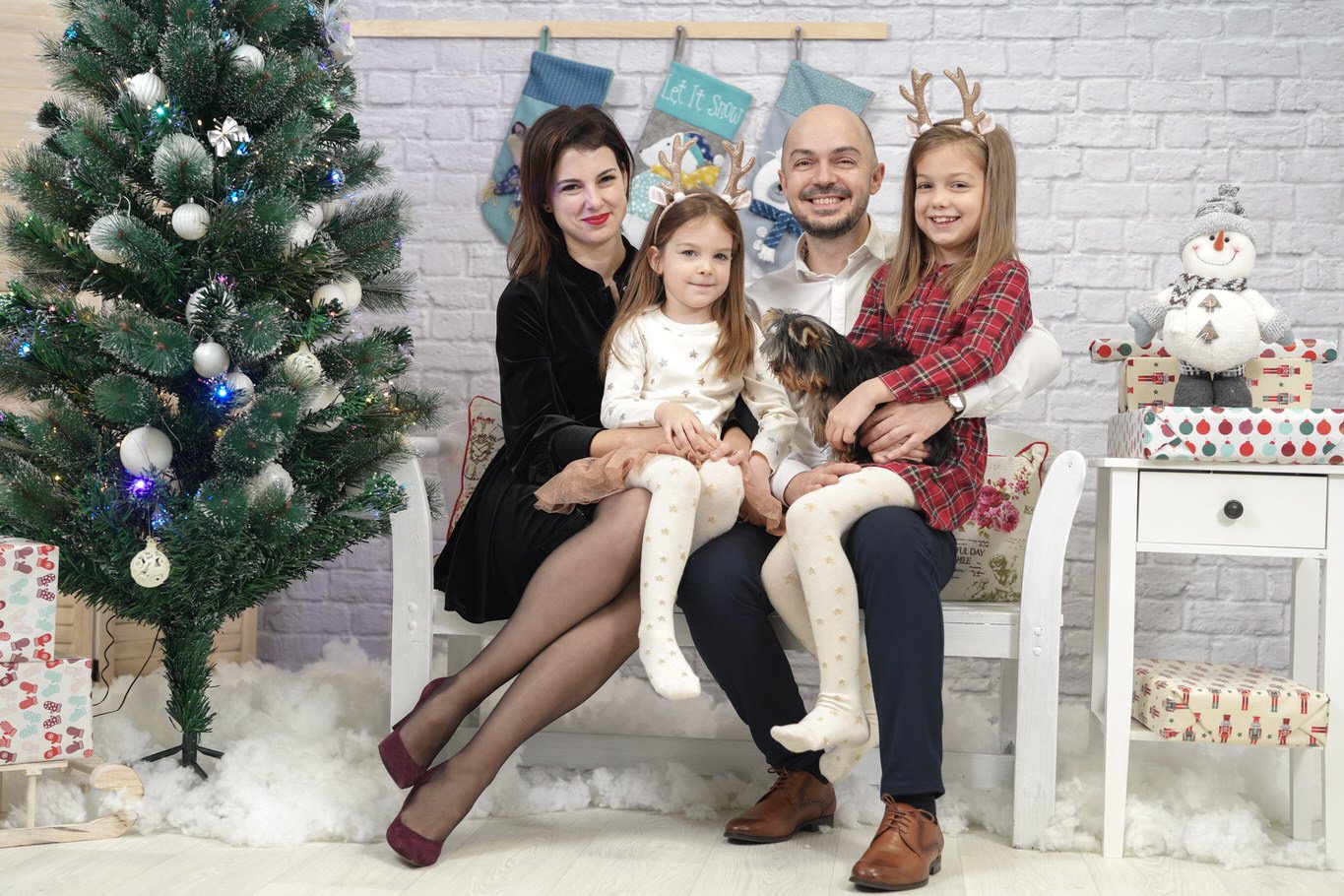 A family of four sitting on a white bench in front of a decorated Christmas tree with ornaments and lights. They are dressed in festive clothing, with two children wearing reindeer antlers. A snowman figurine and Christmas presents are on a table to 