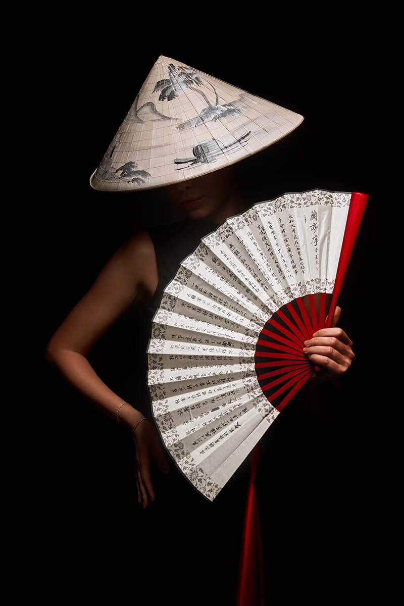 A person partially covered by a large conical Asian hat, holding a decorated folding fan with Chinese calligraphy, against a black background.