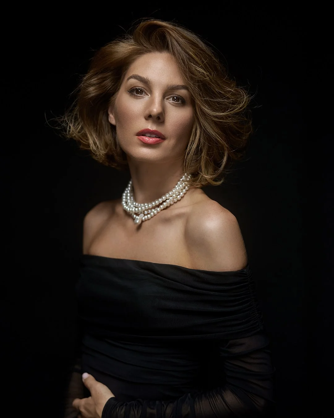 A woman with short, wavy, light brown hair against a black background. She is wearing a black off-the-shoulder dress and a layered pearl necklace, looking at the camera with a confident expression.