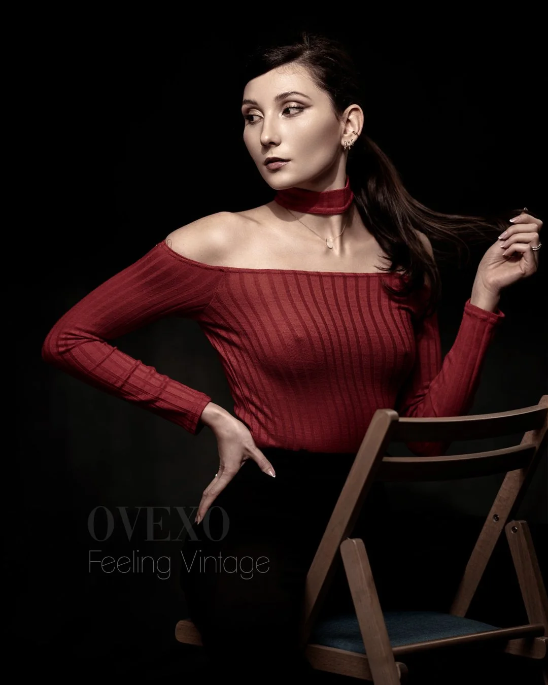 A woman wearing a red off-shoulder top and choker, sitting on a wooden chair against a black background, posing with her hand on her hip and holding her hair, with a neutral expression.