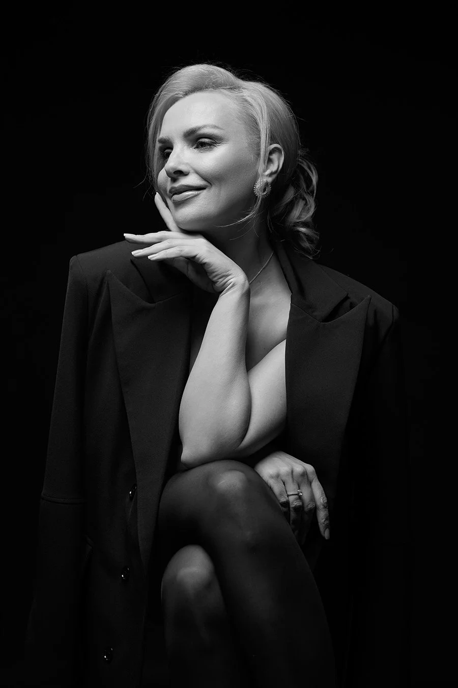 Black and white portrait of a woman with short hair, wearing a suit jacket, with her hand resting on her chin, smiling slightly, against a dark background.