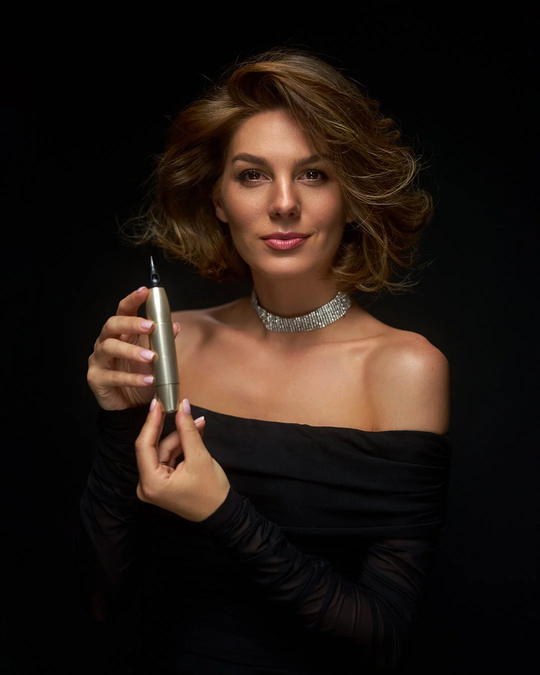 A woman with wavy brown hair, wearing an off-shoulder black dress, a rhinestone choker, and holding a pointing device, standing against a black background.
