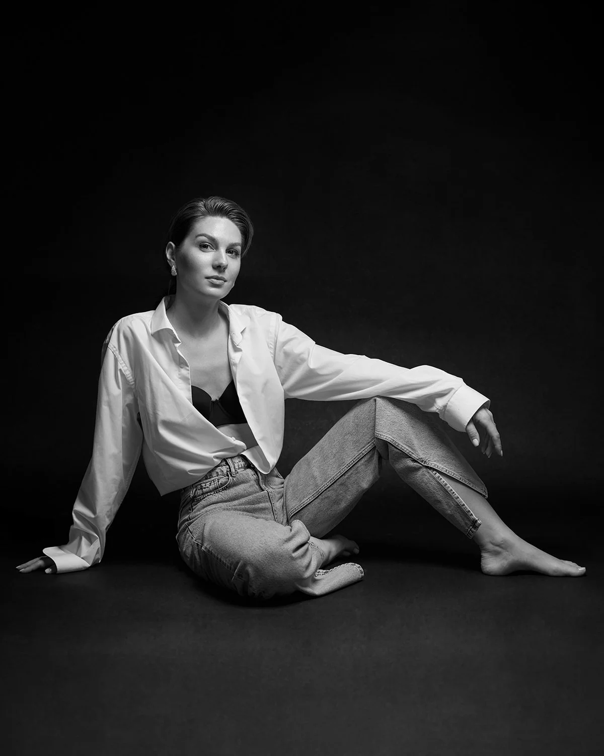 Black and white photo of a woman sitting on the floor, dressed in a white shirt and jeans, with a confident expression.