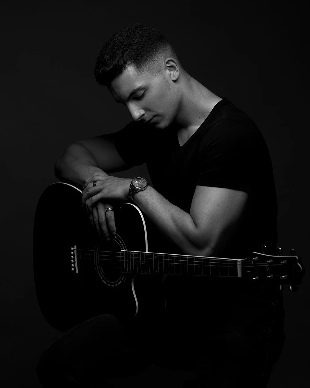 Black and white photo of a young man with short hair, wearing a black t-shirt, sitting with his head bowed, holding an acoustic guitar. He is wearing a watch and rings on his fingers. The background is dark.