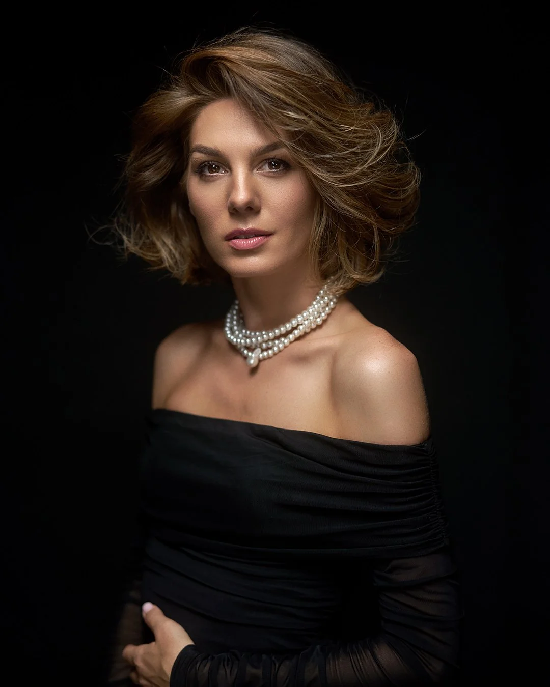 A woman with wavy, shoulder-length brown hair wearing a black off-shoulder dress and pearl necklace, posed against a black background.