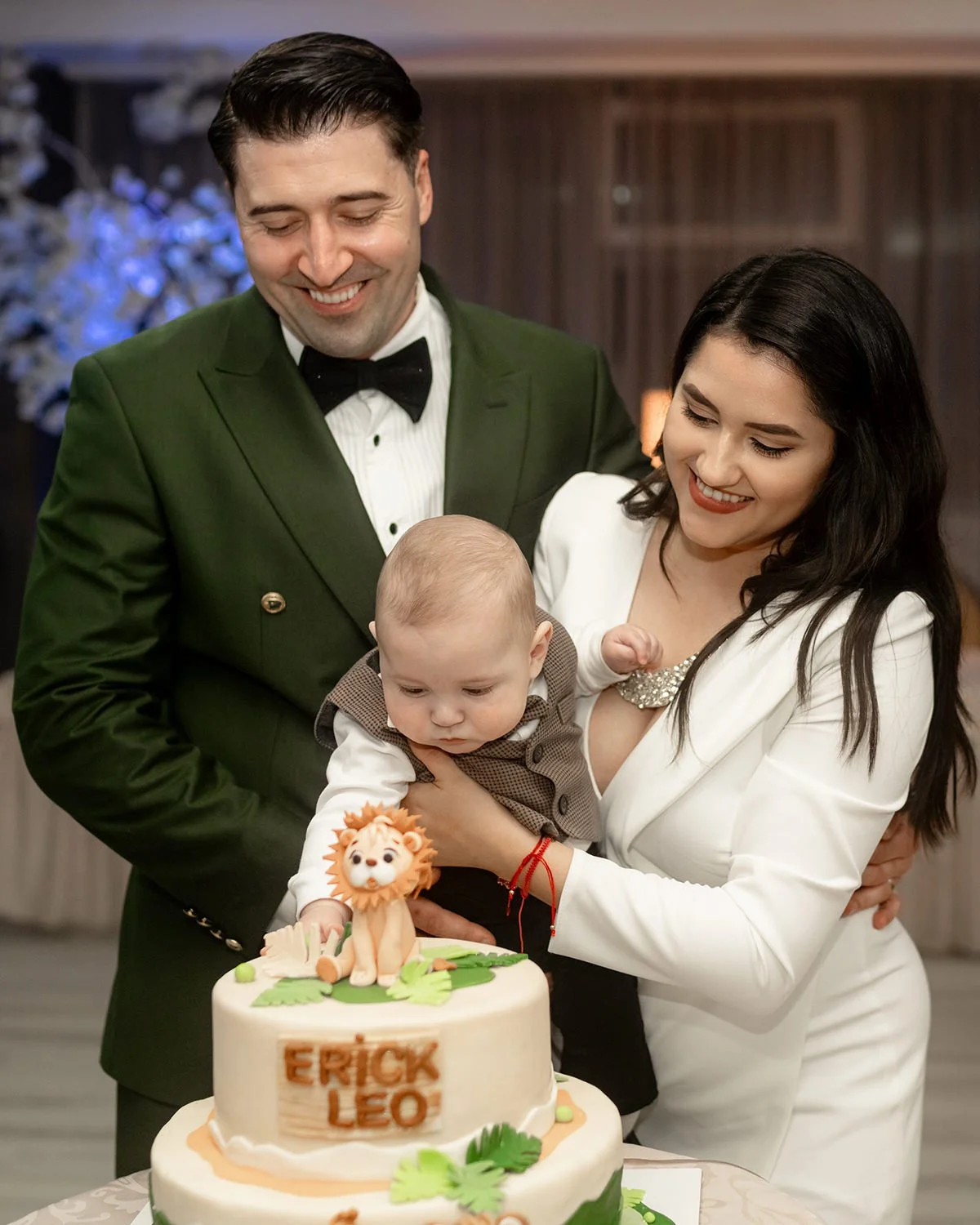 A family celebrating a child's birthday with a cake featuring a lion topper. The father, dressed in a green tuxedo, and the mother, in a white outfit, hold the baby who is reaching towards the cake. The cake has the words 'Erick Leo' on it.