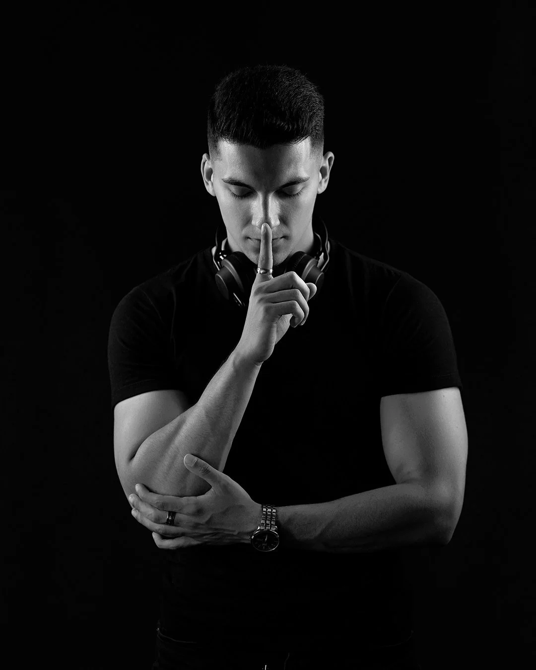 Black and white photo of a young man with a tattooed arm, wearing a black t-shirt, headphones around his neck, and a watch, holding his arm with one hand and placing his finger on his lips.