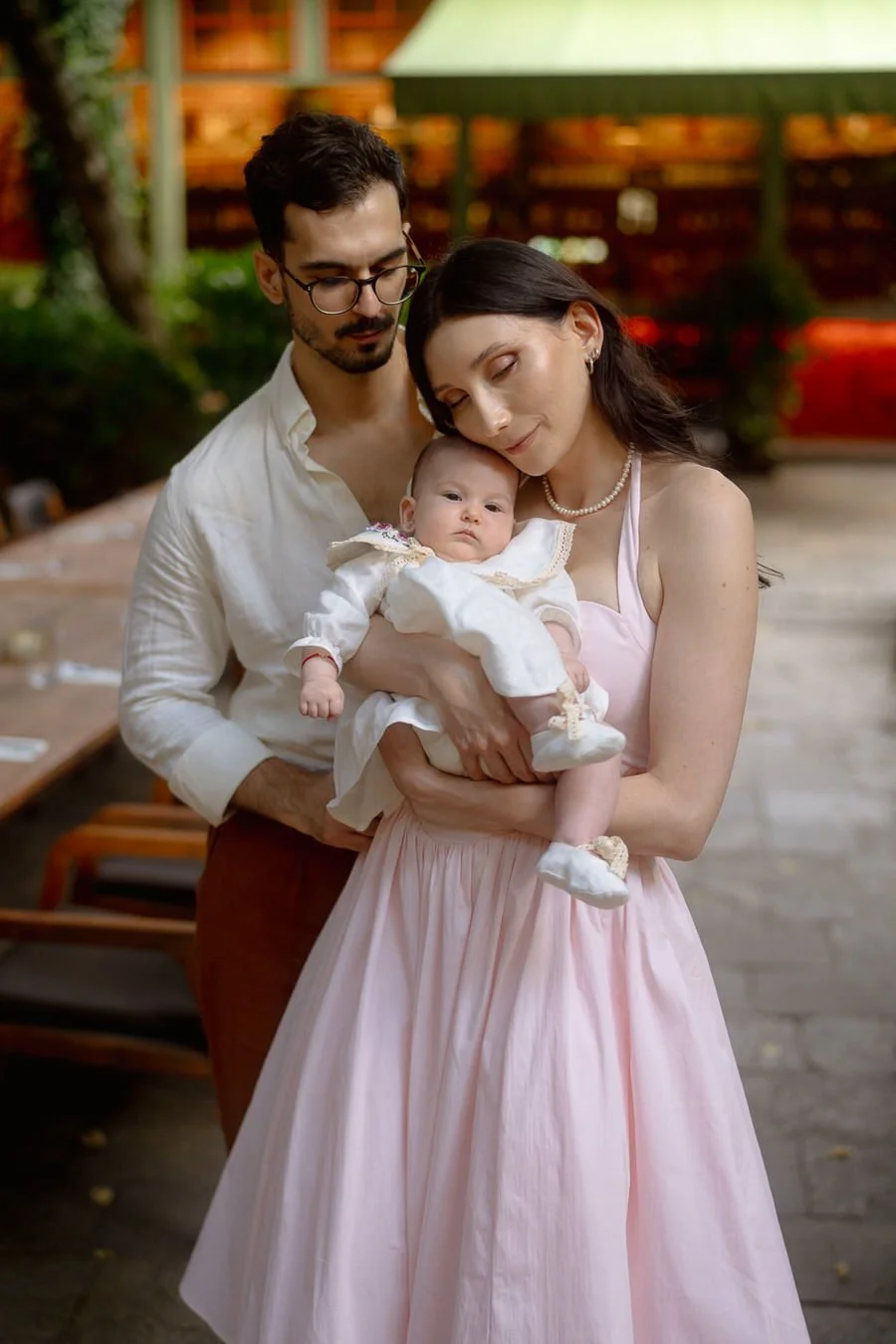 A woman holding a baby dressed in white, with a man standing beside them outdoors, with a green and wooden background.