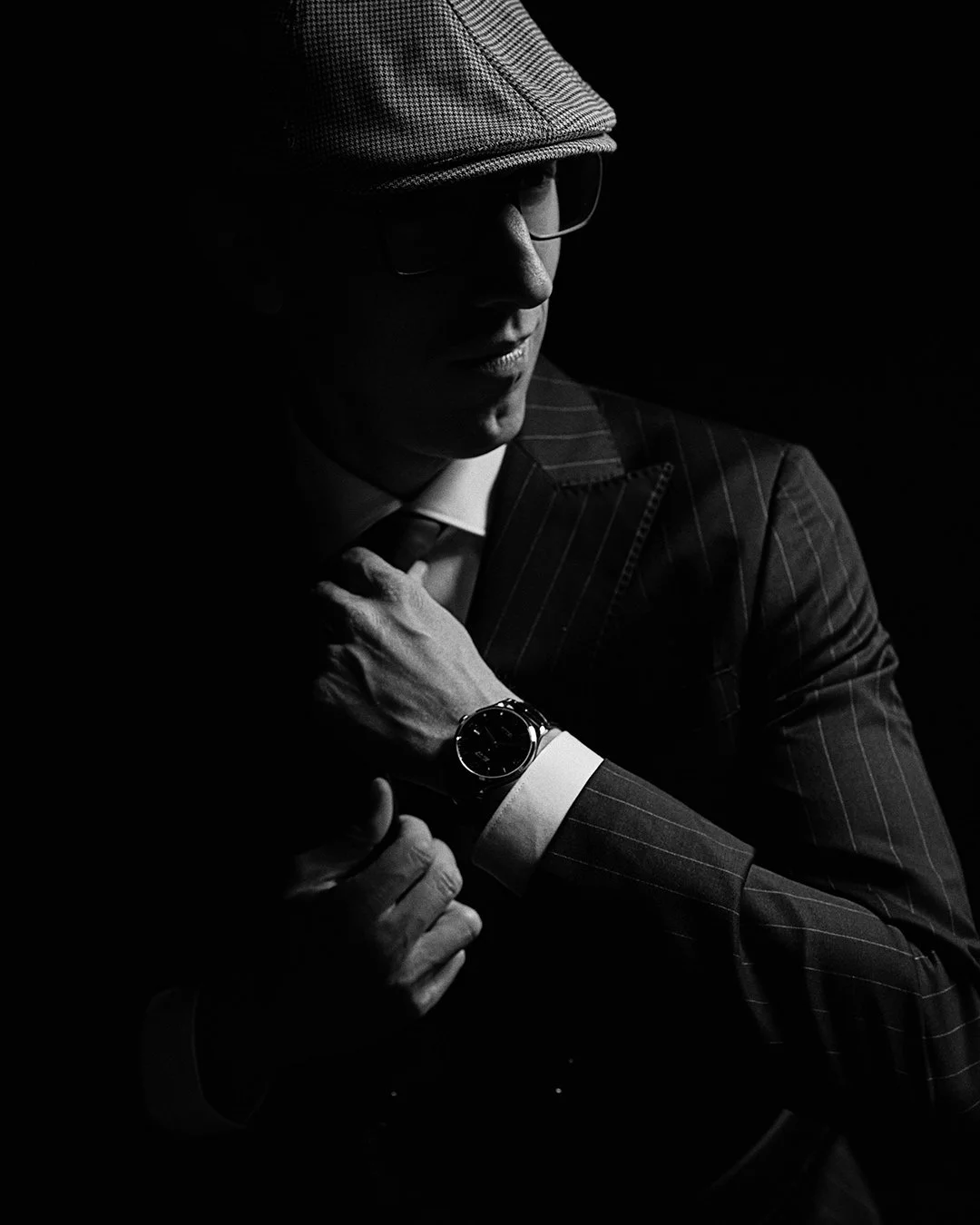 A man in a pinstripe suit wearing a watch, undershirt, bow tie, glasses, and a cap, adjusting his suit in a dark setting.