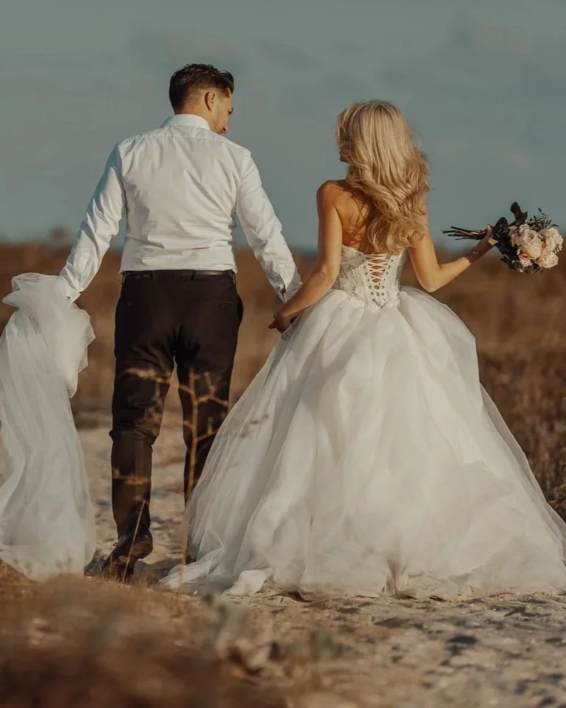 A bride and groom walking hand in hand outdoors, with the bride in a white wedding gown holding a bouquet, and the groom in a white shirt and dark pants, on a sandy area with open sky.