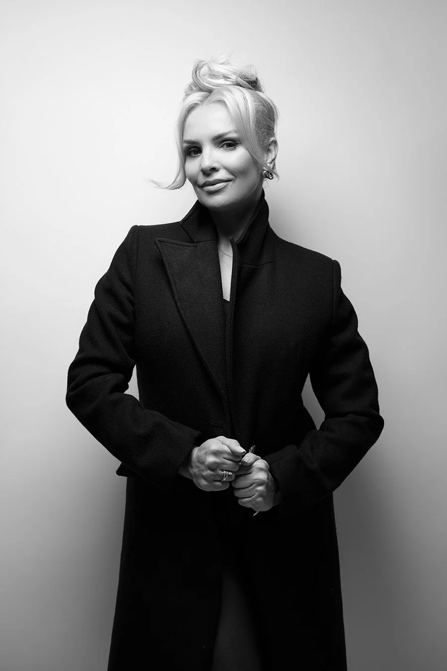Black and white portrait of a woman with short blonde hair styled in an updo, wearing a dark coat, standing against a plain light background.