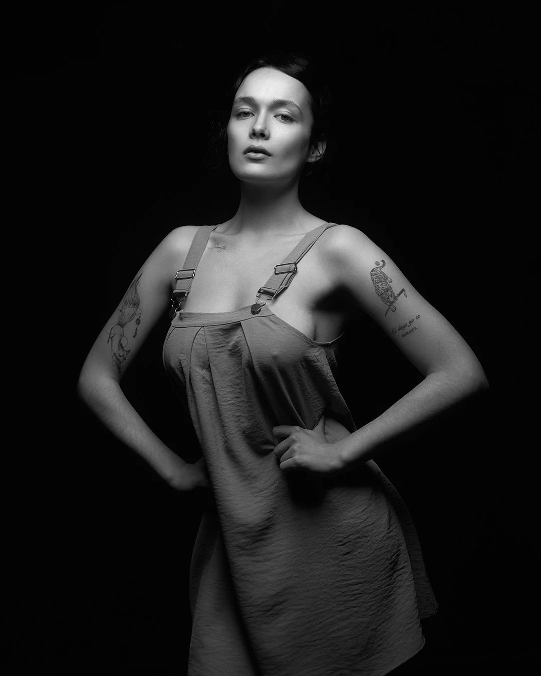 Femeie cu tatuaje pe brațe pozând cu mâinile în șolduri într-un studio foto, purtând o rochie cu bretele, iluminare dramatică pe fundal negru.