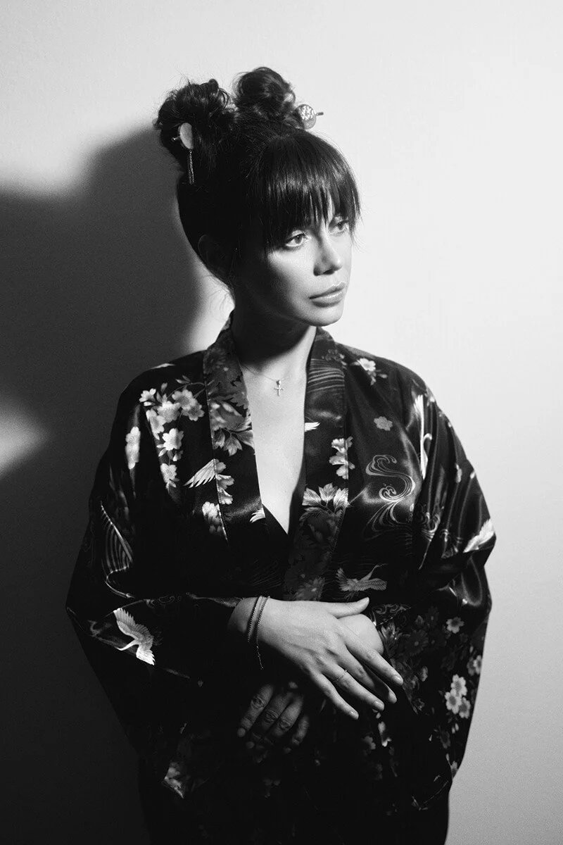 Black and white portrait of a woman with dark hair styled in an updo with bangs, wearing a floral kimono and a delicate cross necklace, standing against a plain wall with a shadow cast on her left side.