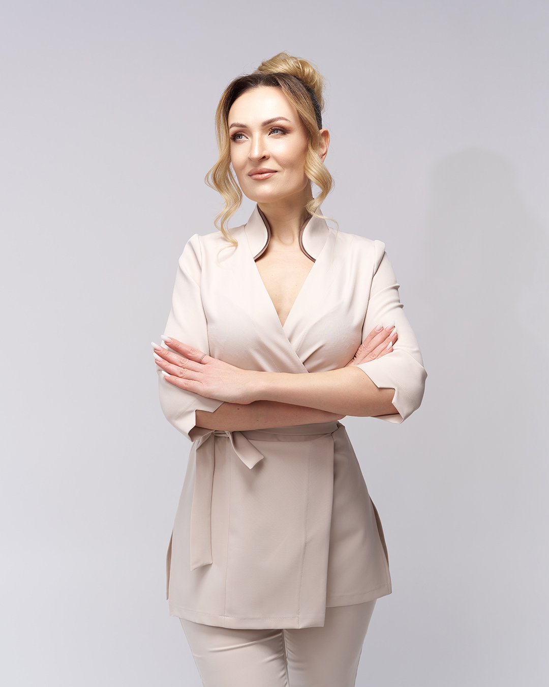 A woman with blonde hair styled in loose curls, wearing a beige wrap blazer and pants, standing with her arms crossed against a plain light gray background.