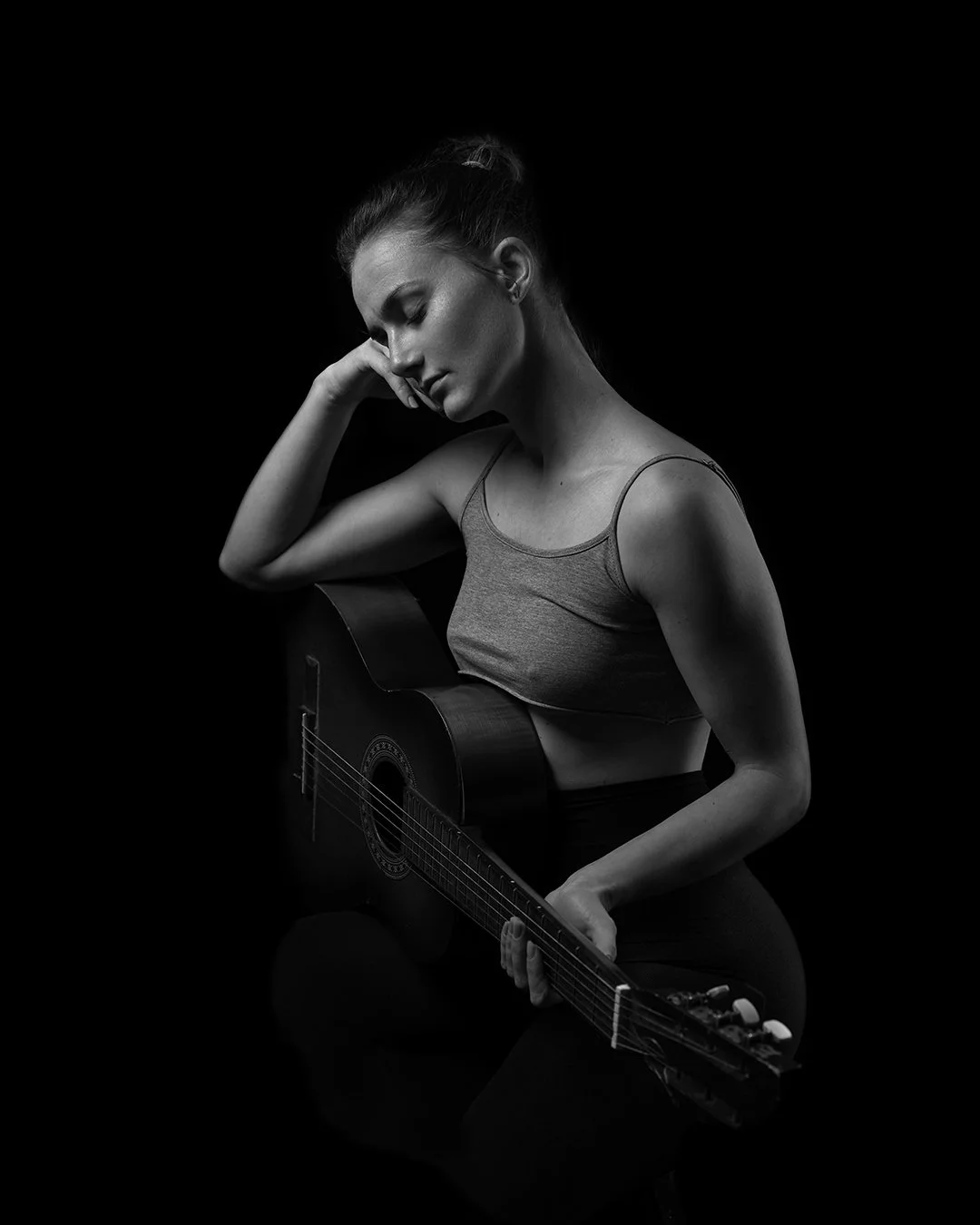 A black and white photo of a woman holding a guitar, with her eyes closed and a contemplative expression, against a dark background.