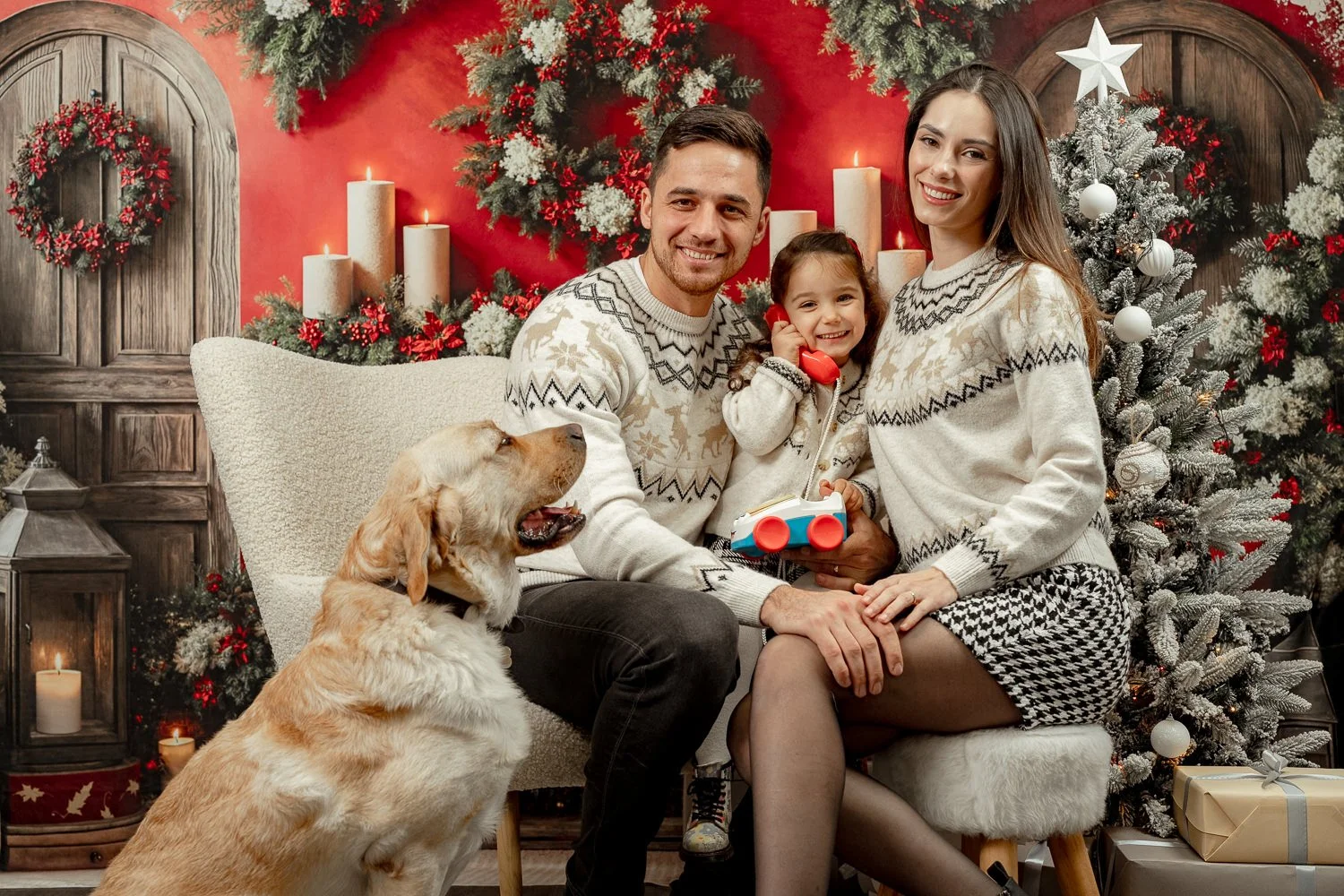 O familie zâmbitoare cu un copil mic și un câine Golden Retriever, pozând într-un studio foto decorat festiv pentru Crăciun, cu brad și fundal roșu cu coronițe.