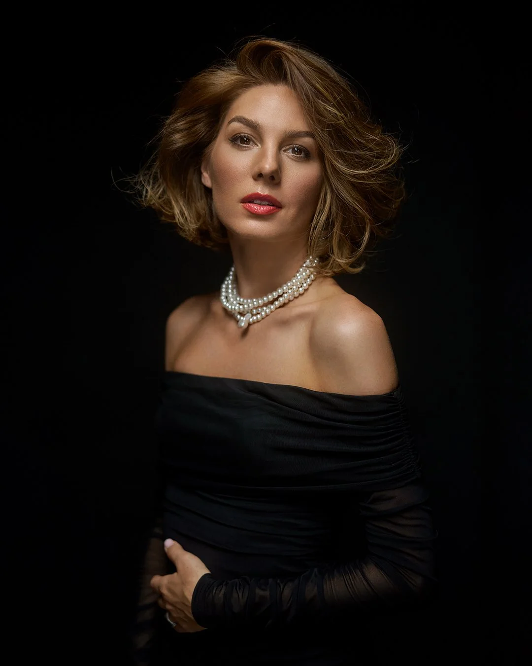 A woman with shoulder-length wavy hair wearing an off-the-shoulder black dress and a layered pearl necklace poses against a dark background.
