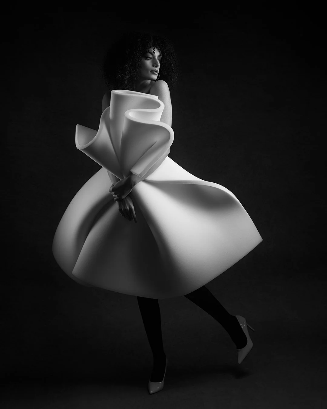 A black and white creative portrait of a woman in an avant-garde dress with large, sculptural folds, standing against a dark background.