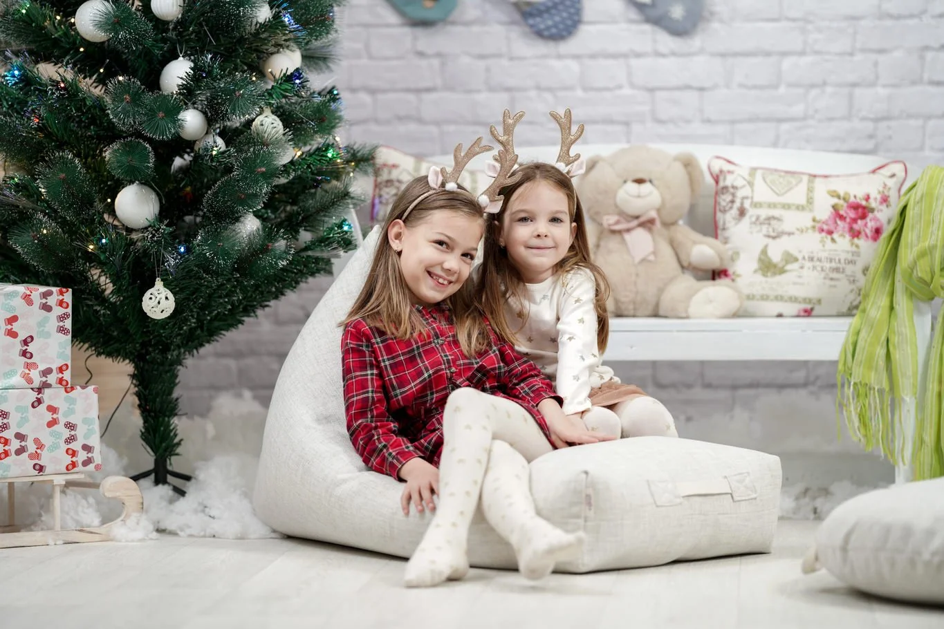 Two young girls sitting on a white bean bag chair in front of a decorated Christmas tree, one wearing reindeer antlers, both smiling.
