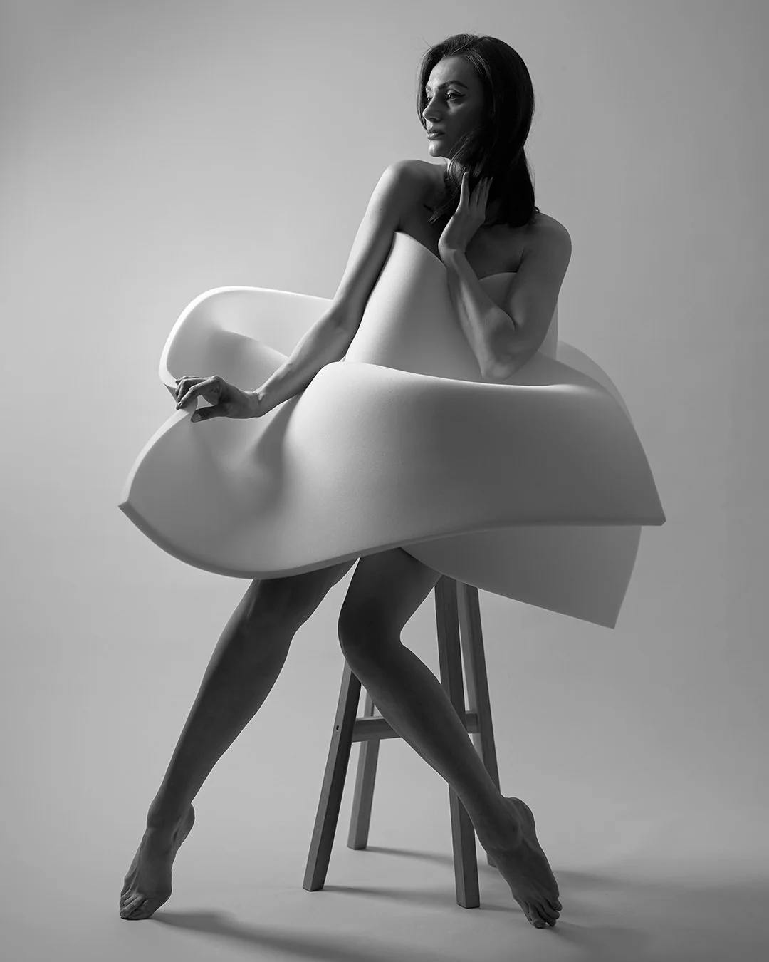 Black and white photograph of a woman sitting on a stool with a large, abstract, sculptural piece encircling her. She poses with her left hand gently touching her neck, and her right hand on the sculpture, with her legs crossed. The composition empha