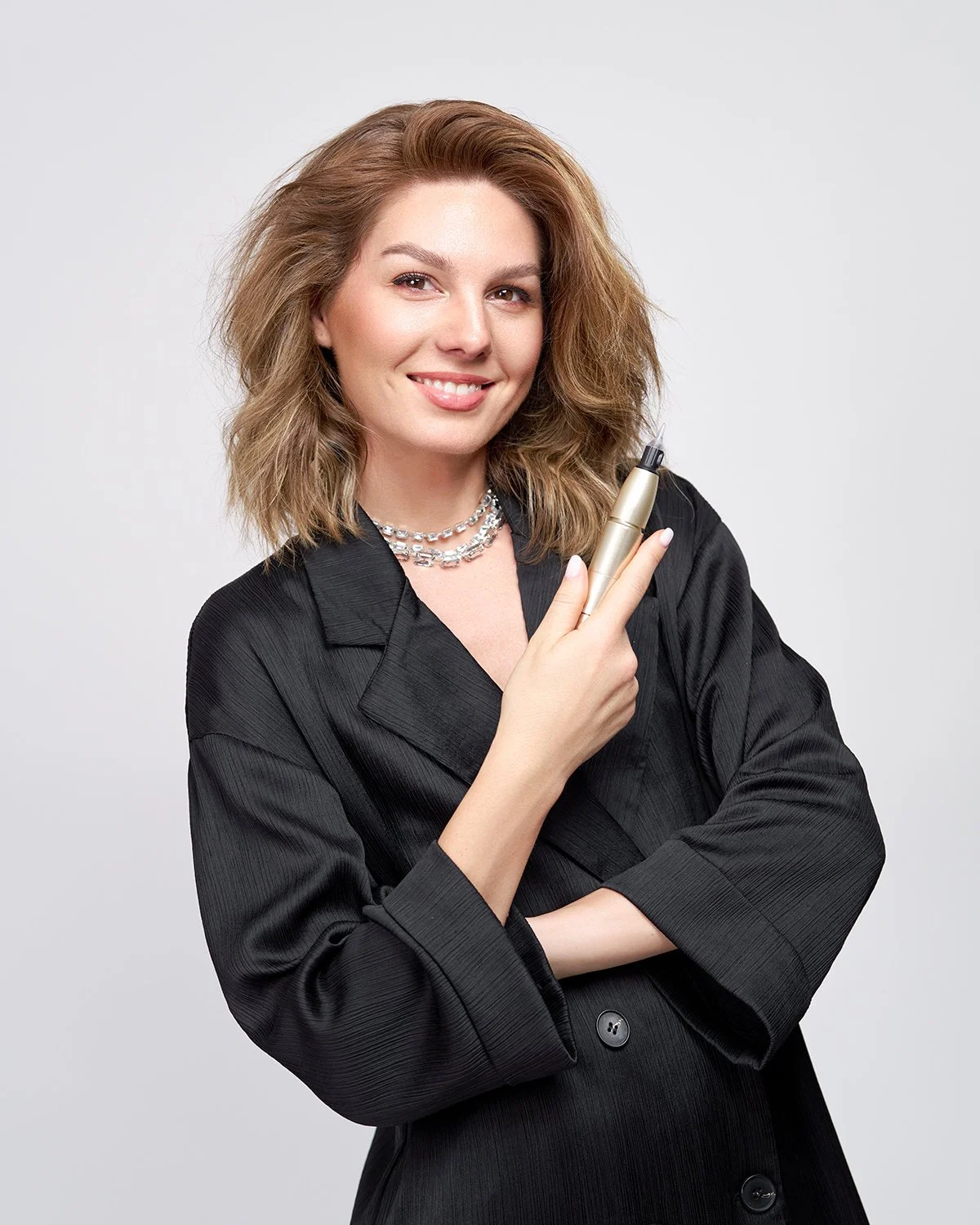 A woman with shoulder-length wavy hair smiling, wearing a black blazer, holding a gold-colored pen, and wearing a layered pearl necklace.