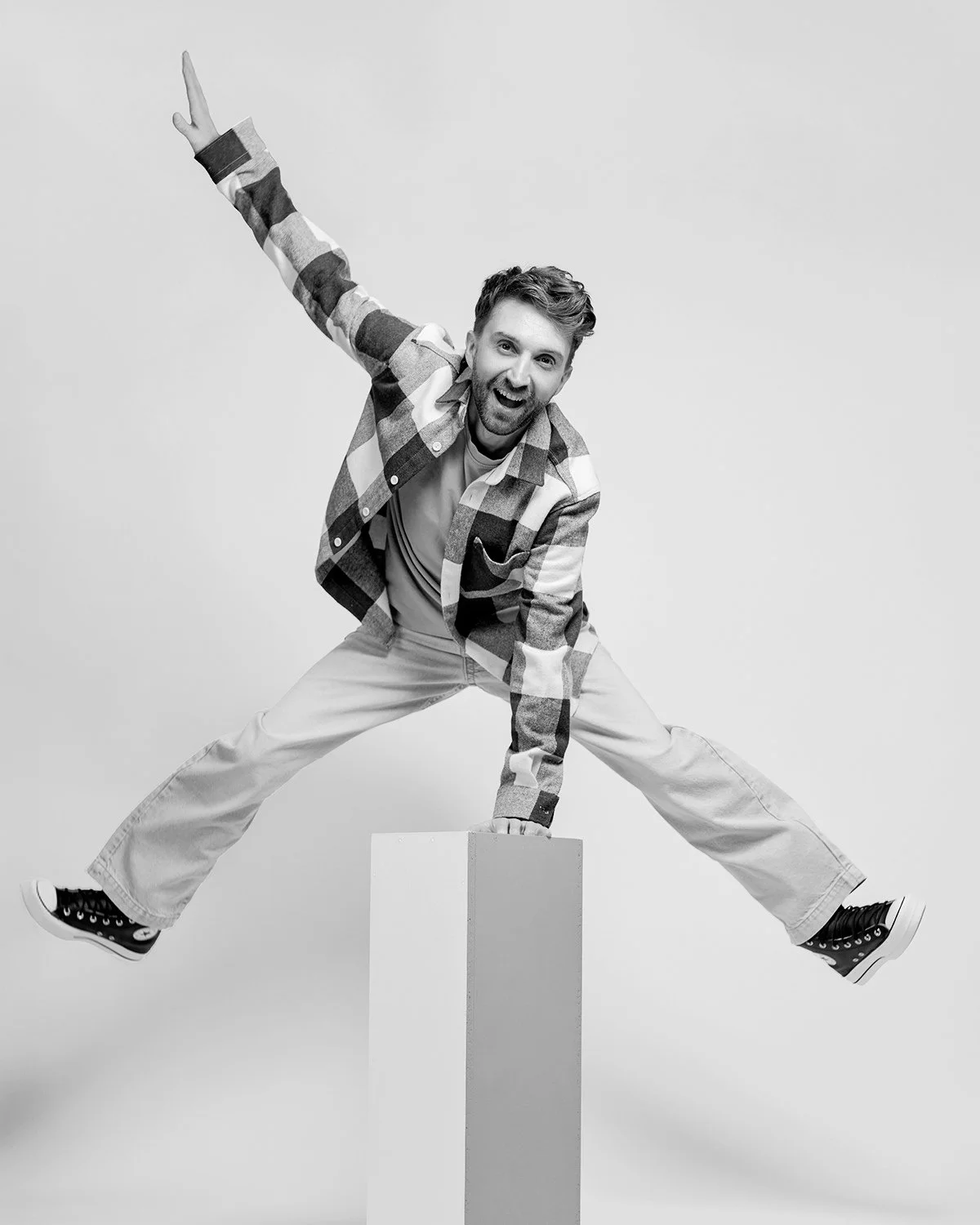 A young man is balancing with one hand on a pedestal, legs wide apart, smiling, in a studio with a plain background.