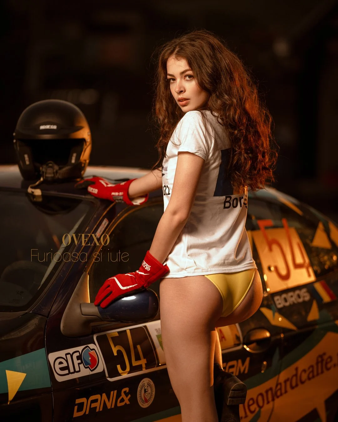 A woman with long curly hair posing next to a racing car, wearing a white racing shirt, yellow underwear, red racing gloves, and black boots, with a black helmet on the car's roof.