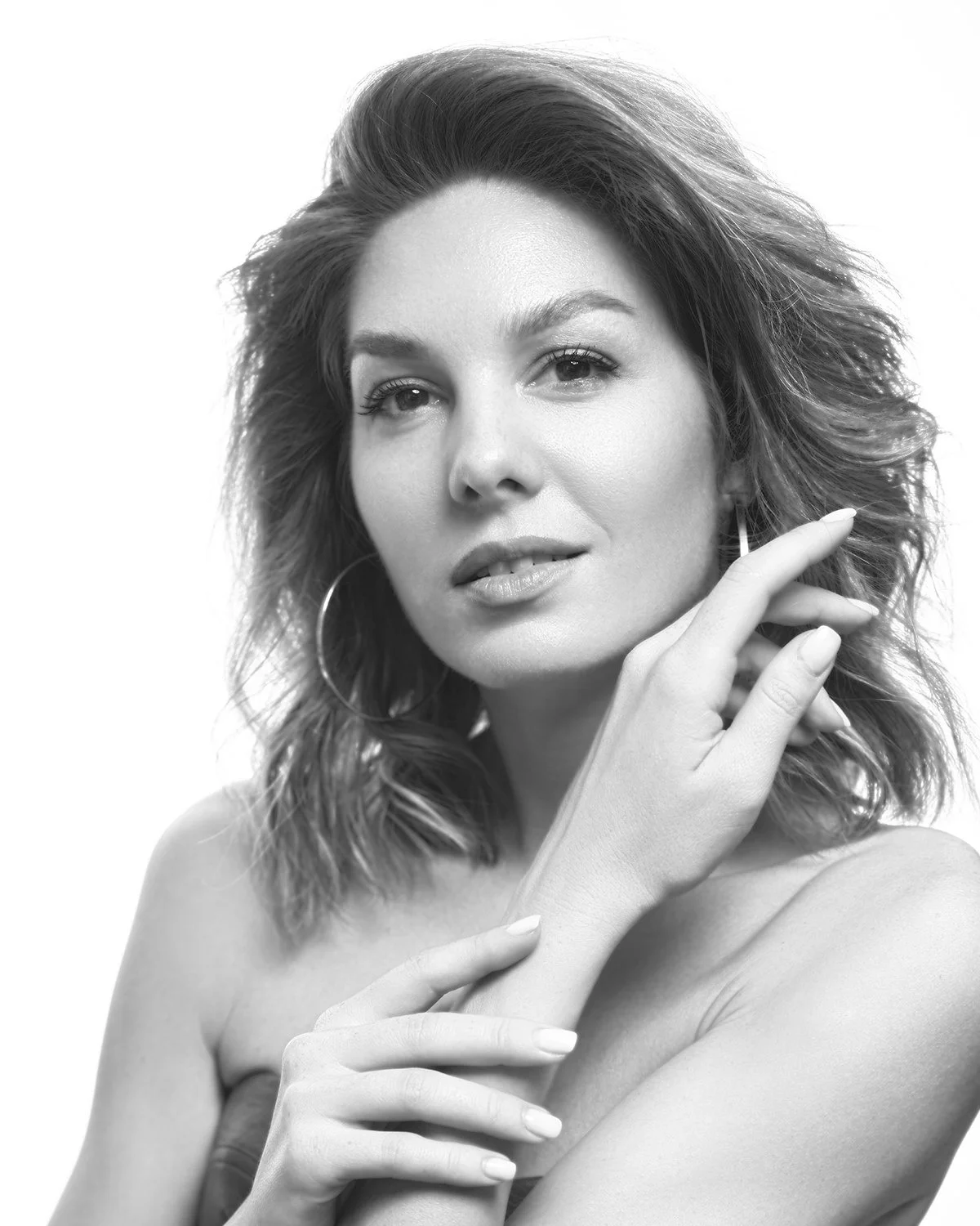 Black and white portrait of a woman with wavy hair, wearing hoop earrings, looking at the camera with a slight smile, one hand near her face, and the other on her shoulder.