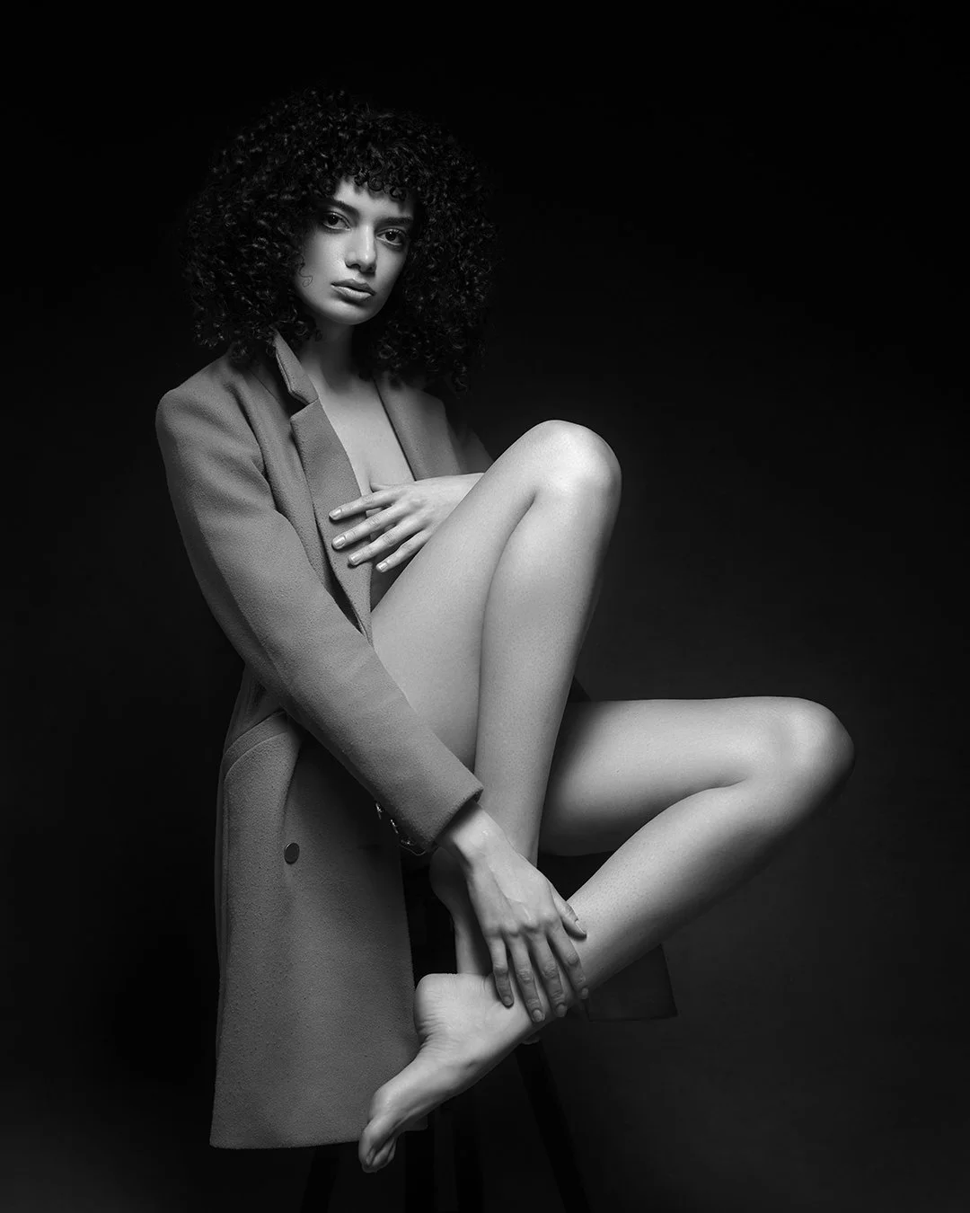 Black and white photo of a woman with curly hair, wearing a coat, sitting on a chair with one leg bent and crossed over the other, covering her chest with one hand, against a dark background.