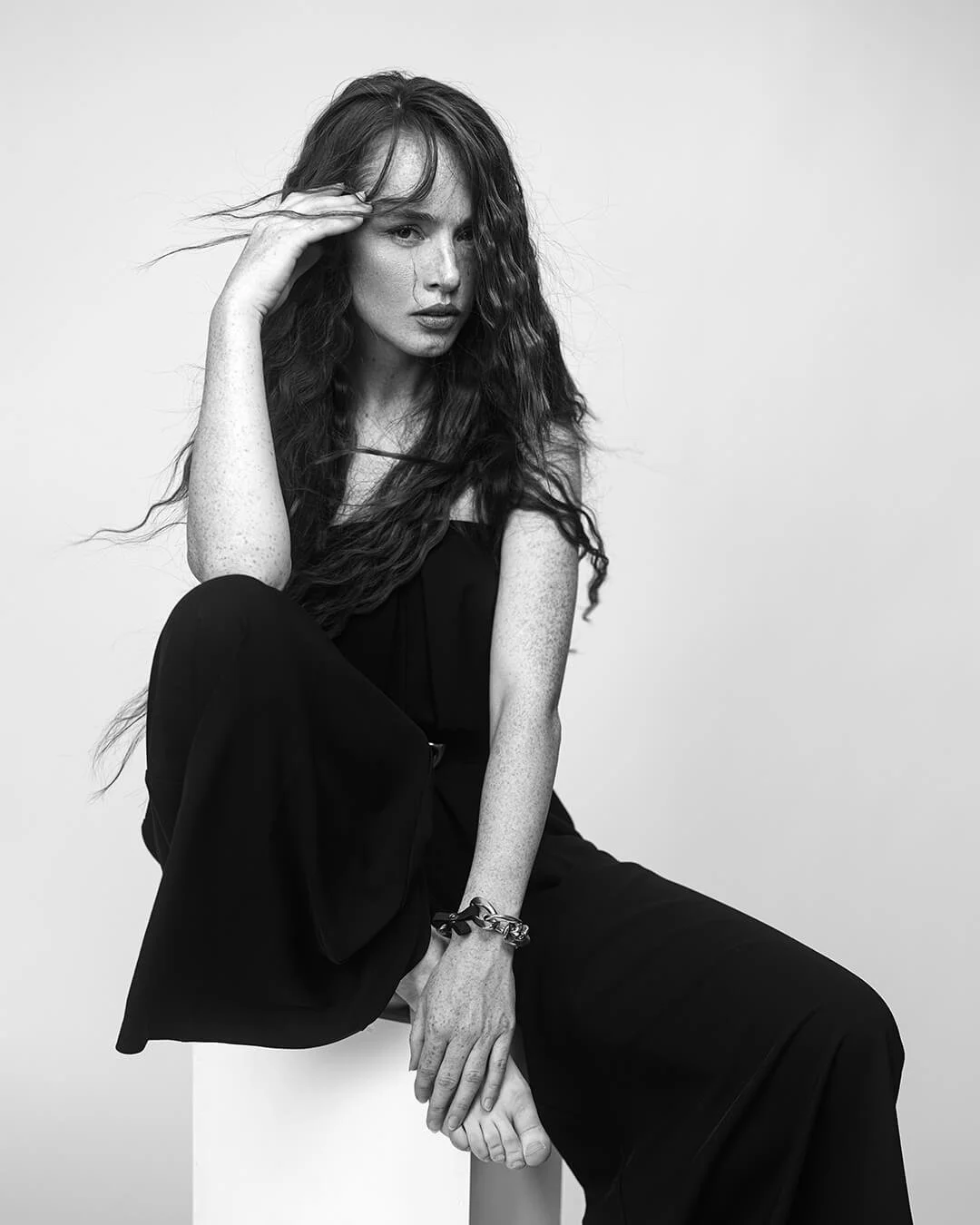 Black and white photo of a woman with long wavy hair, sitting with one knee up, wearing black clothing, and touching her head with one hand.