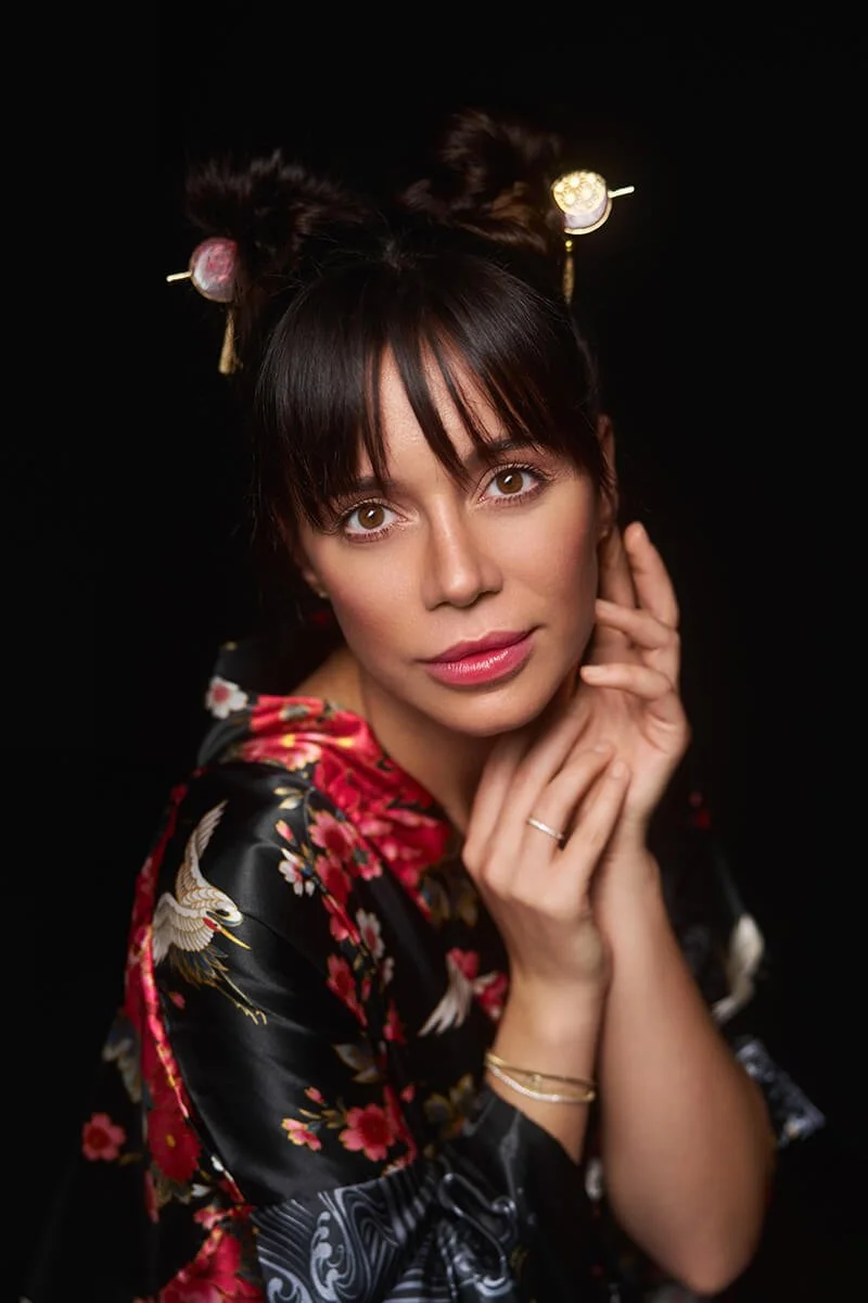 A woman with dark hair styled in two buns with hair ornaments, wearing a black kimono with red and pink floral patterns, against a black background.