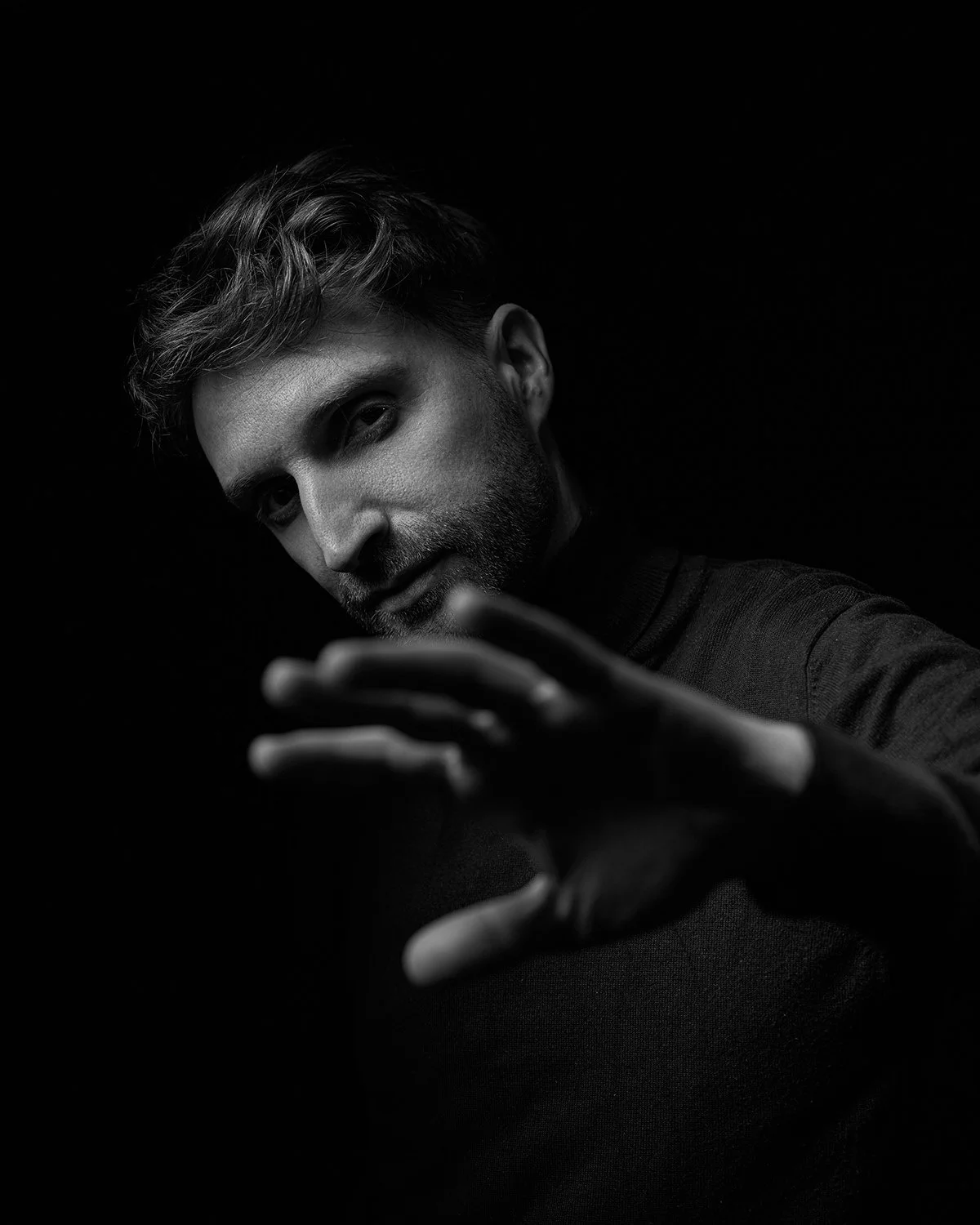 Black and white photo of a man with styled hair and a beard, reaching out with his right hand toward the camera, against a dark background.