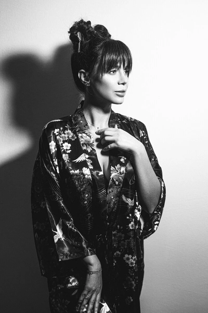 Black and white photograph of a woman wearing a floral kimono, with her hair styled in an elegant updo, standing against a plain white background and looking to the side.