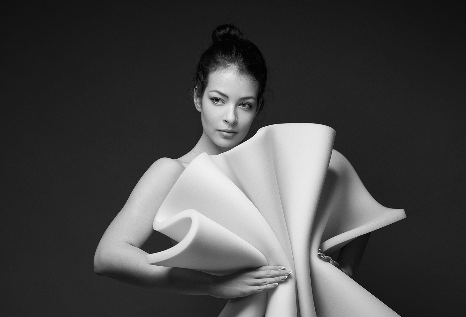 Portret alb-negru de studio cu un model feminin purtând o structură vestimentară albă, sculpturală și avangardistă pe fundal gri închis.